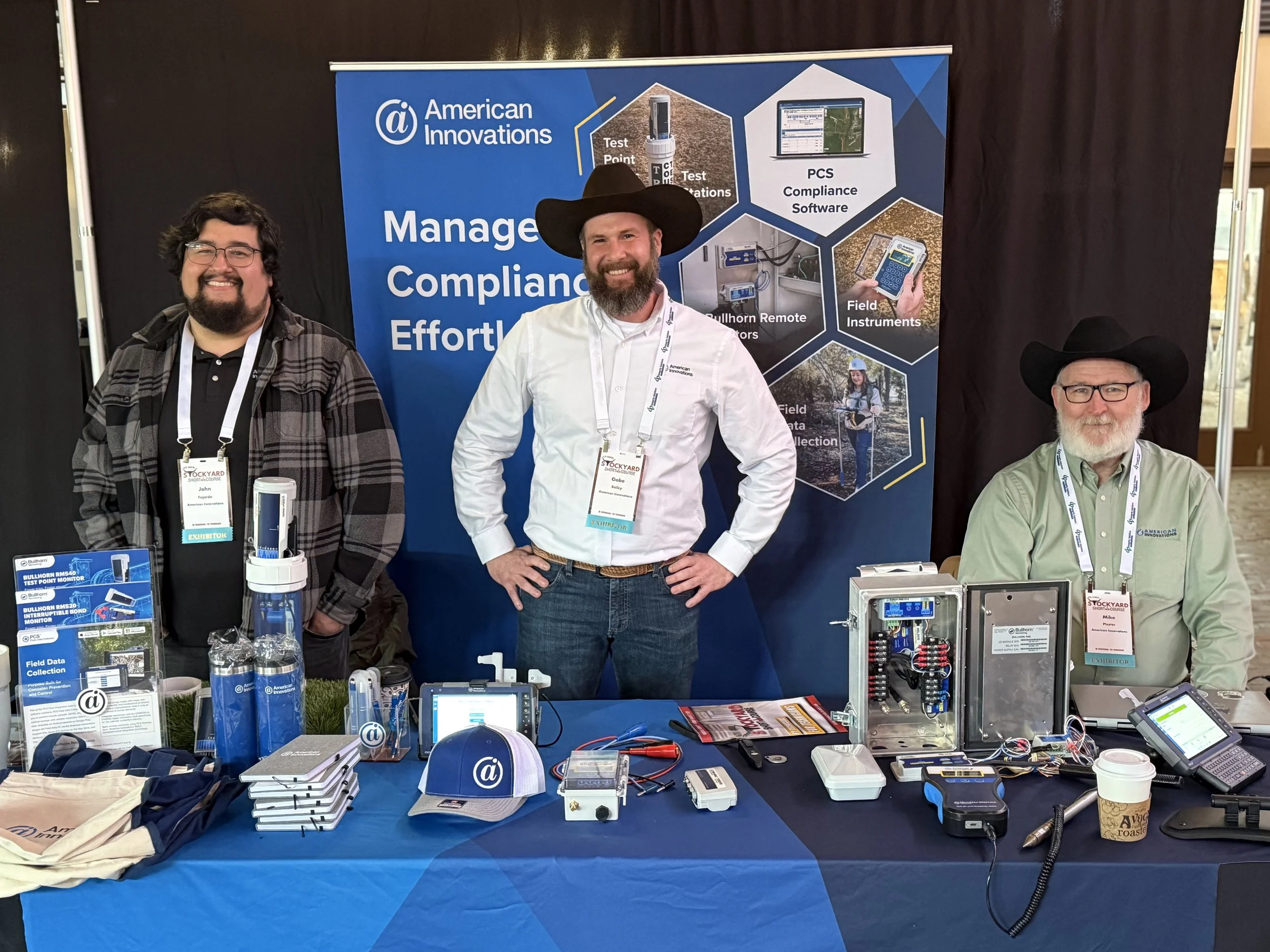 Three men standing behind a booth at a trade show, with equipment and promotional items on the table. The background features a blue banner with information about American Innovations.