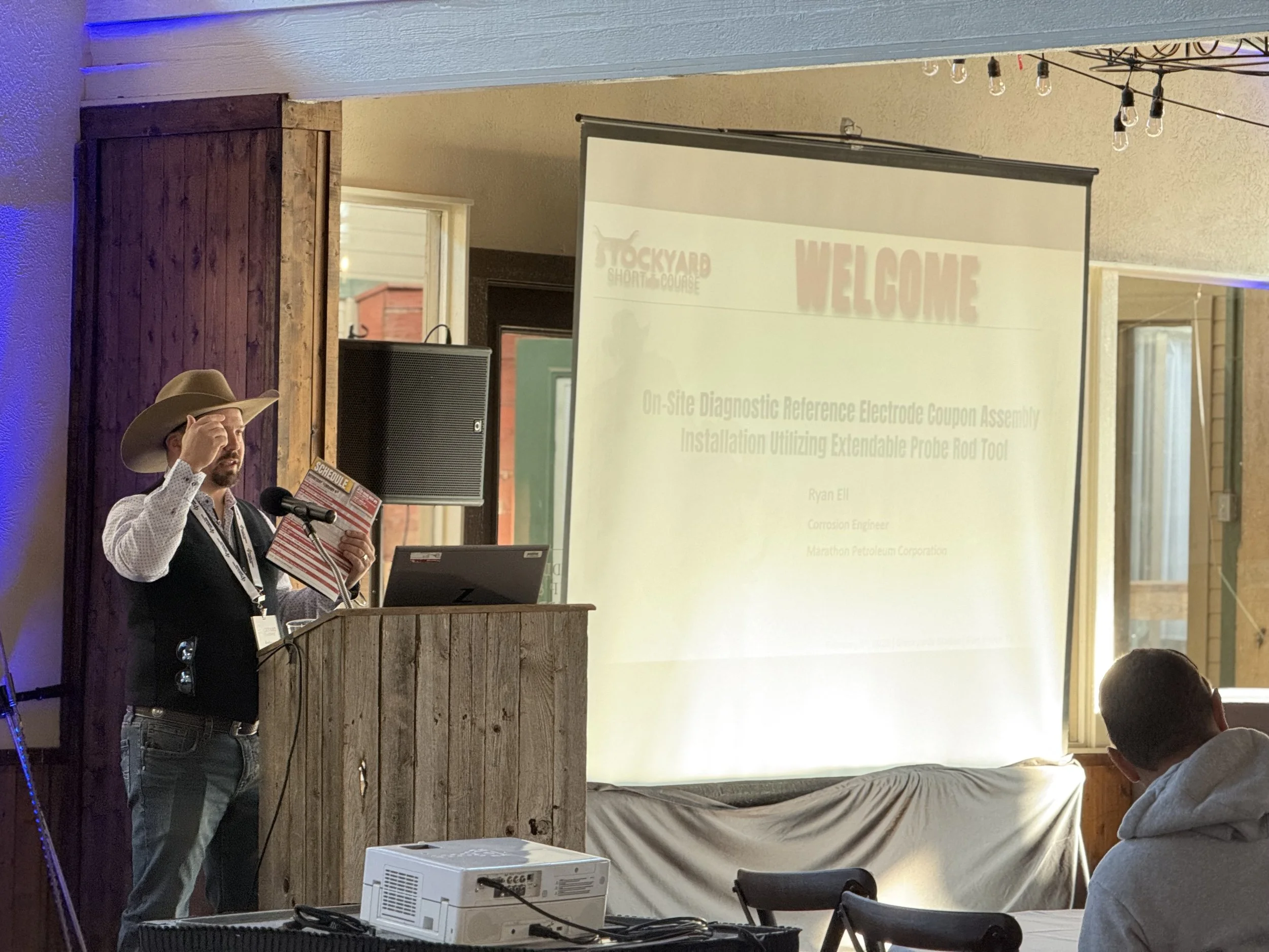 A man in western attire, wearing a cowboy hat, standing at a wooden podium, holding a booklet, giving a presentation in a rustic room with a large projector screen displaying a presentation slide.