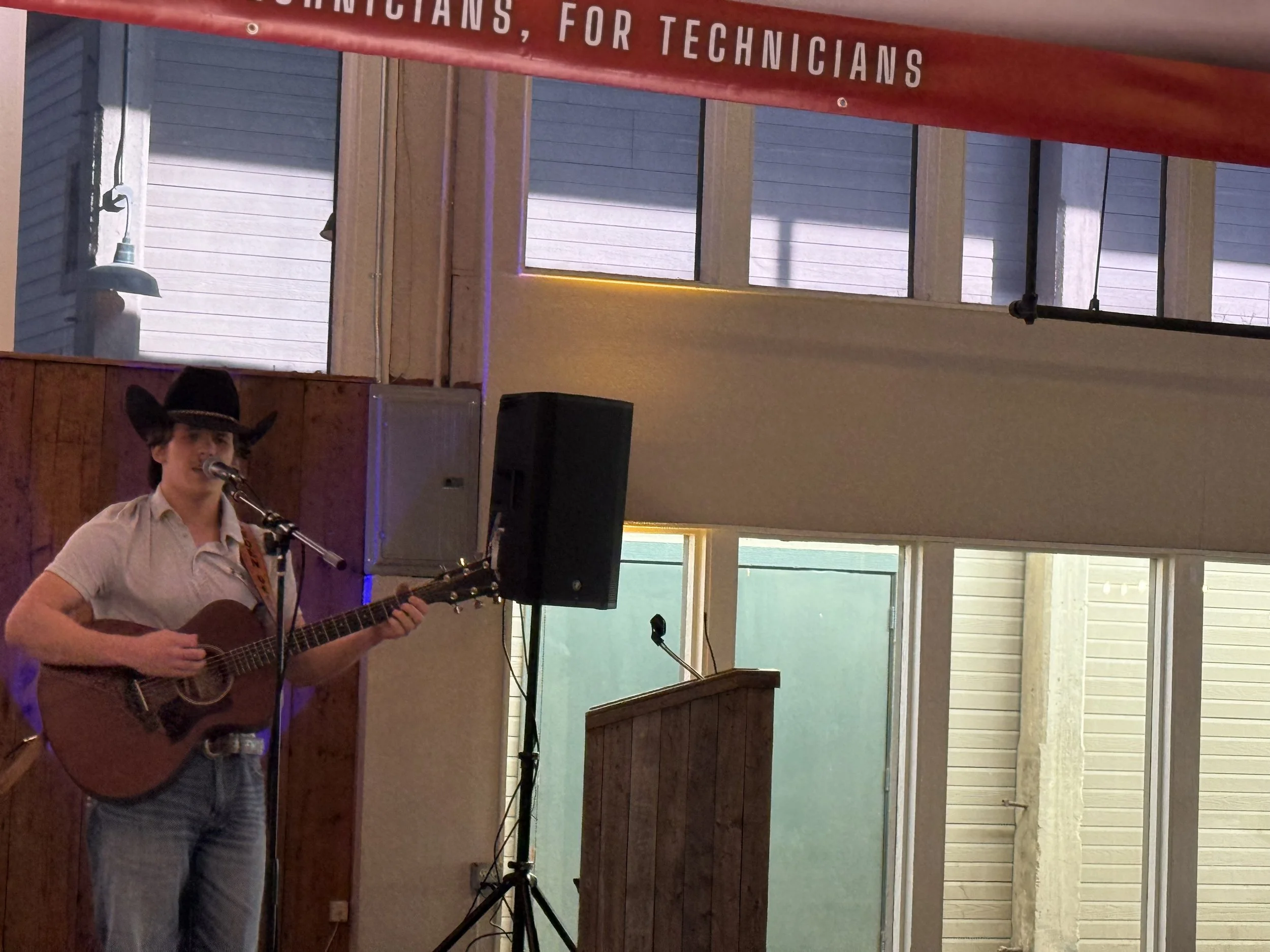 A person wearing a cowboy hat and shirt playing an acoustic guitar and singing into a microphone indoors, with speakers and a wooden podium nearby, and large windows in the background.