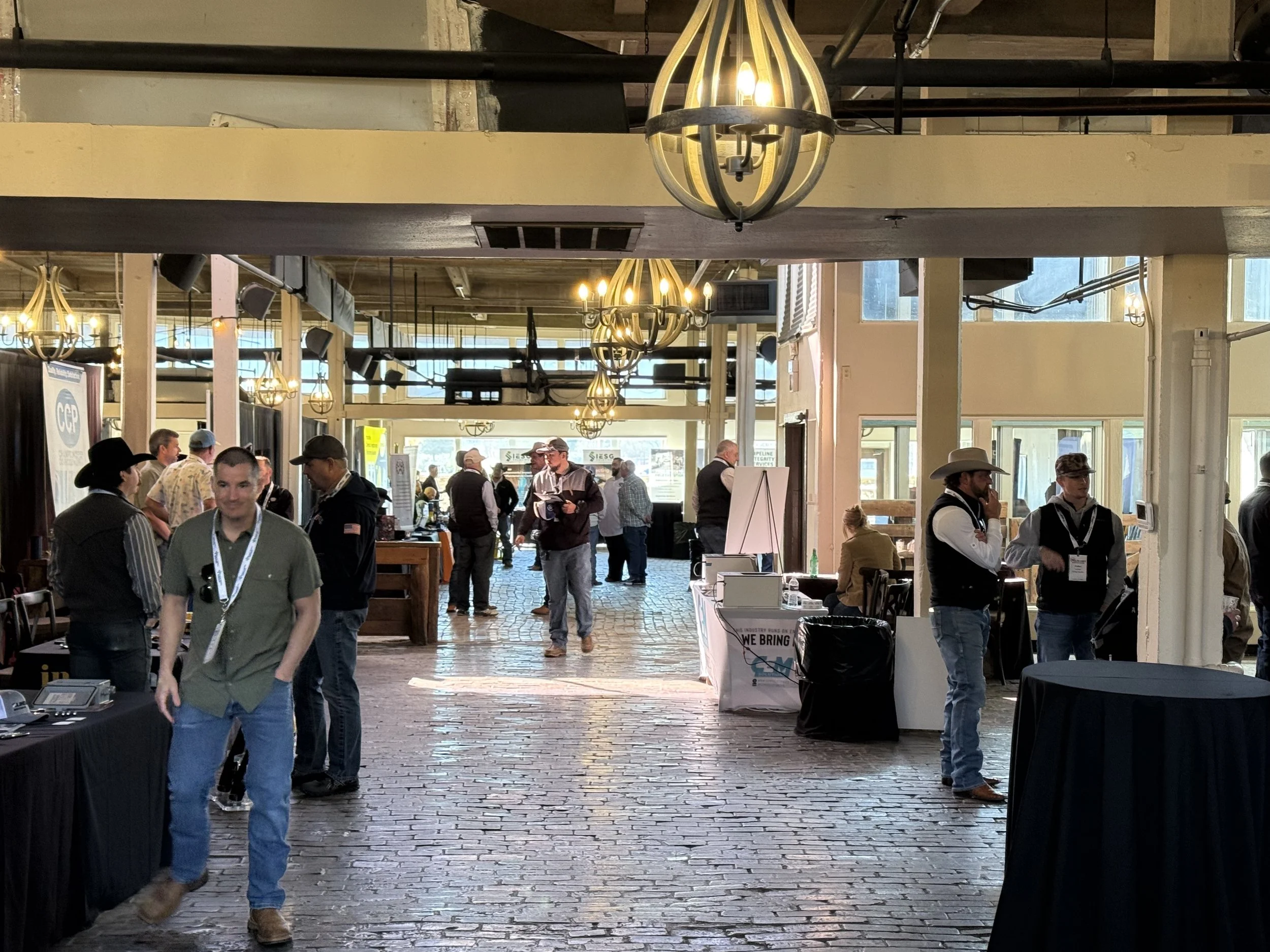 People attending an indoor event or conference, walking around and talking at various booths and tables, with large hanging chandelier lights overhead and natural light coming through windows.