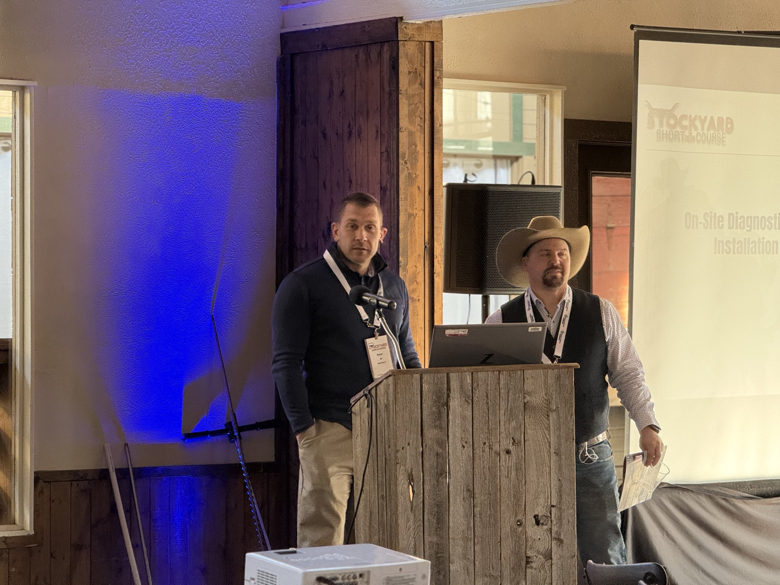 Two men standing at a presentation podium during a conference or seminar in a rustic venue, with a large screen to the right displaying a presentation slide and a window behind them showing a brick building outside.