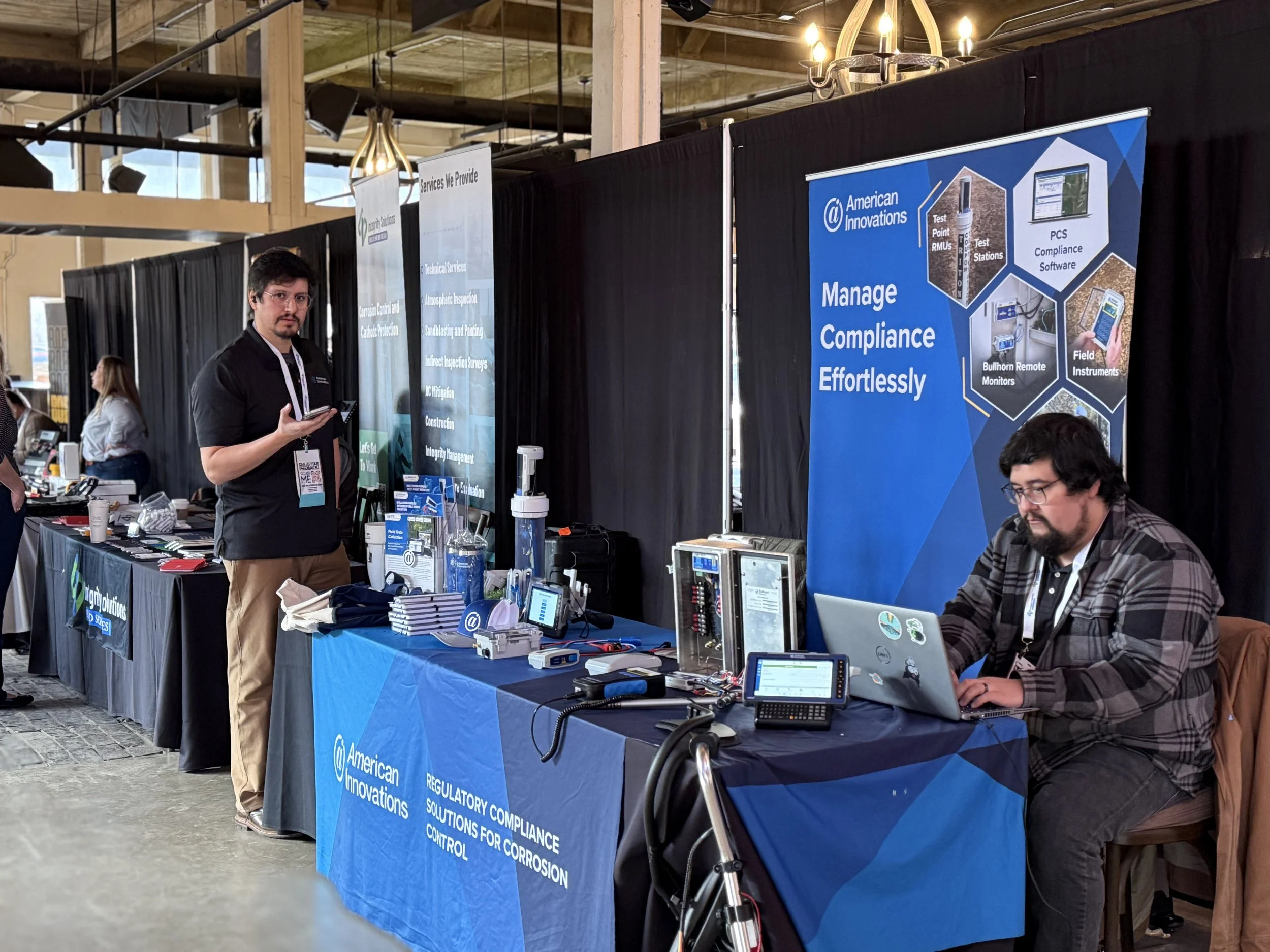 At a trade show booth for American Innovations, two men are present; one standing and holding a tablet, the other sitting at the booth working on a laptop. The booth features a large blue banner with the company logo and information about managing co