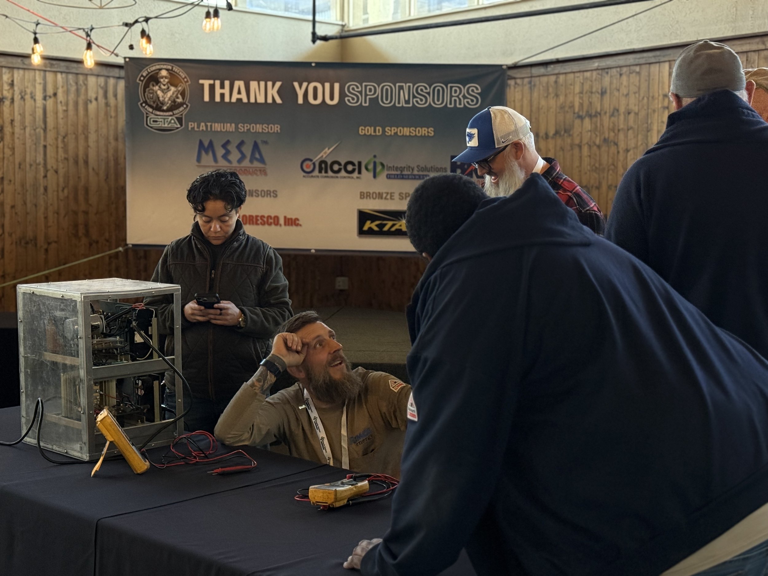 People gathered around a man seated at a table, engaged in conversation; a woman stands nearby looking at her phone; a large banner in the background thanks sponsors; the setting is indoors with string lights overhead.