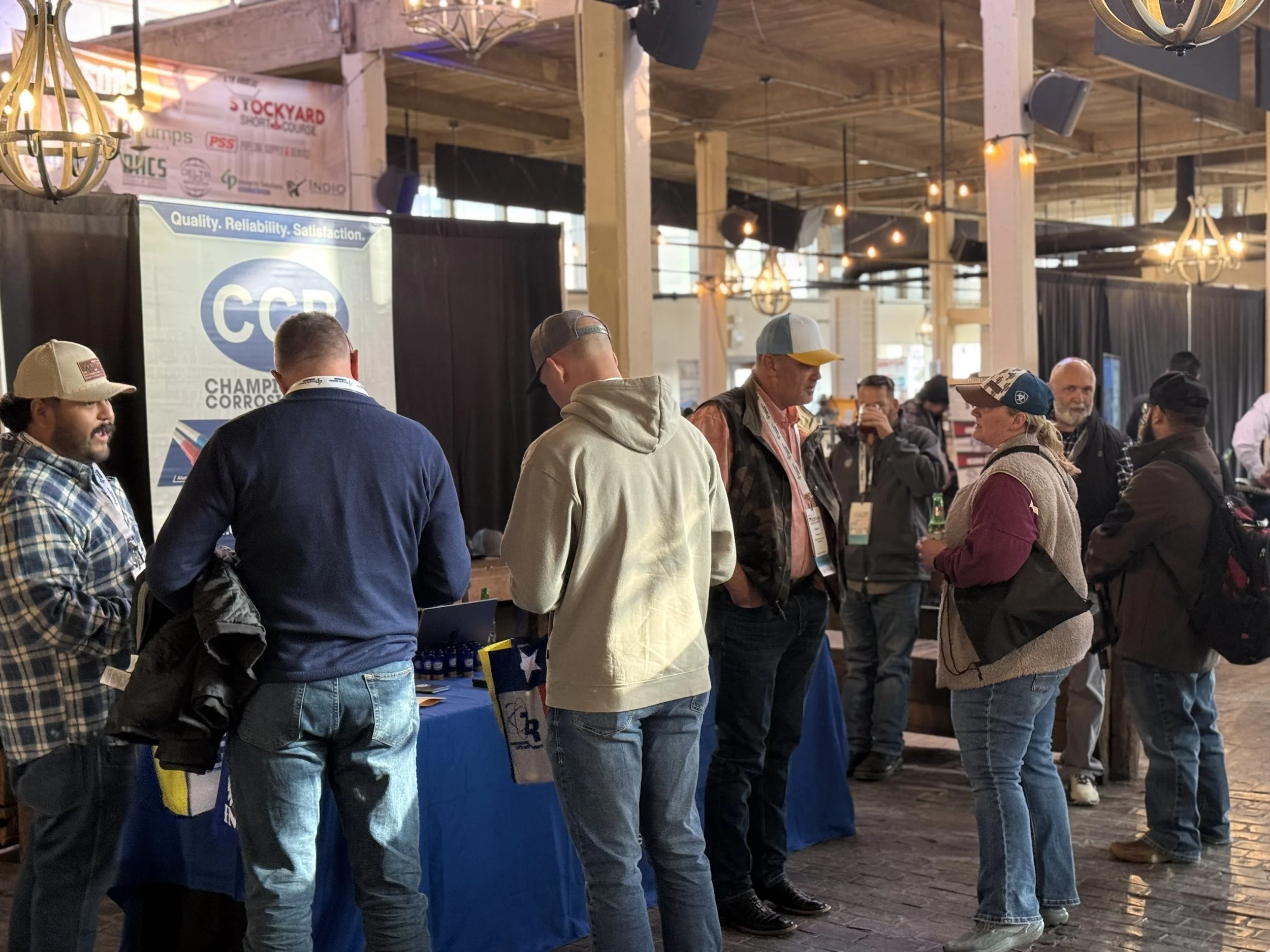 People attending an indoor trade show or conference, standing in line or gathering around a booth with a CCP (Championship Corrosion) banner. The setting features wooden beams, hanging lights, and various exhibit tables.