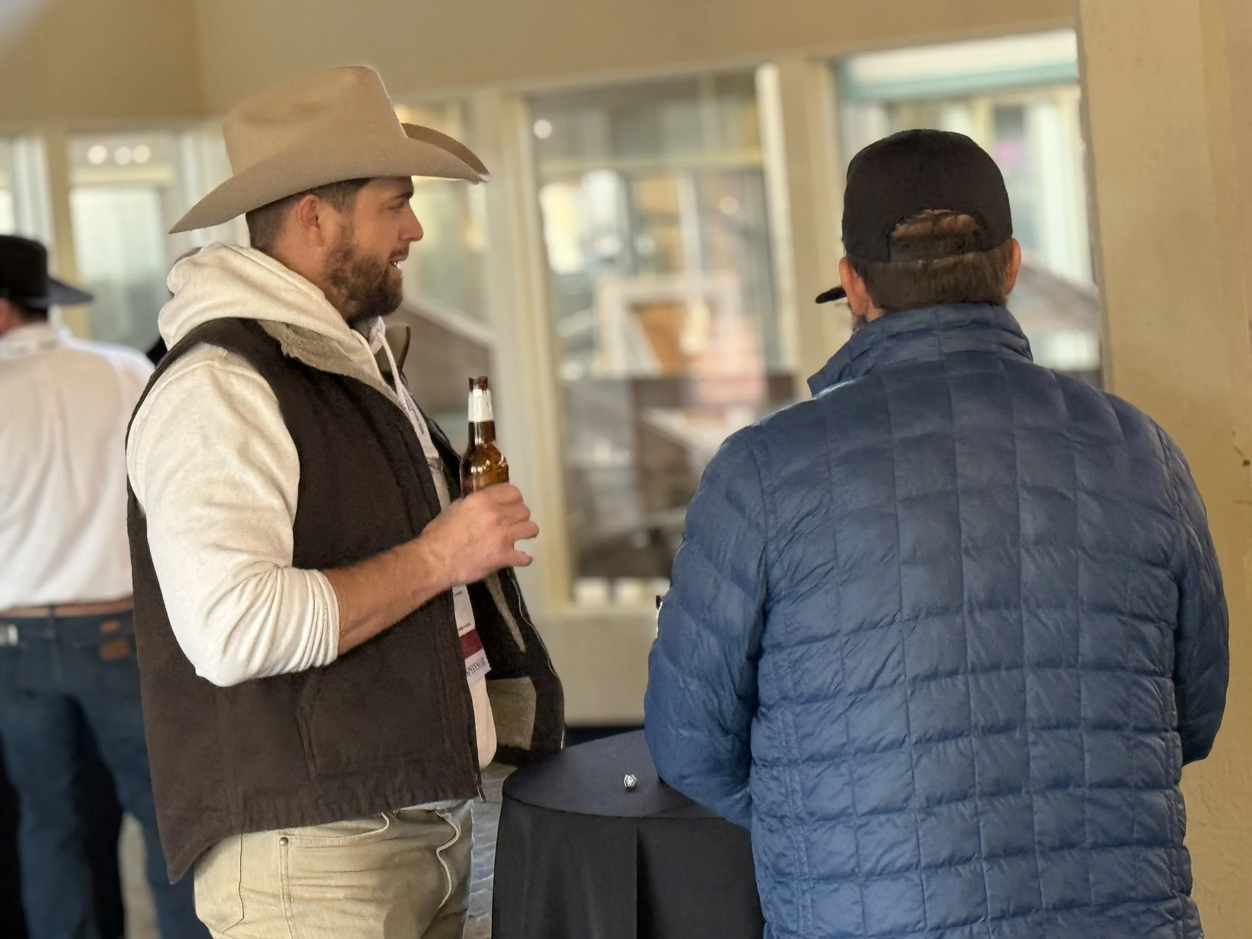 Two men socializing indoors, one wearing a cowboy hat and holding a beer, the other in a baseball cap with a quilted jacket, near a table with a black tablecloth.
