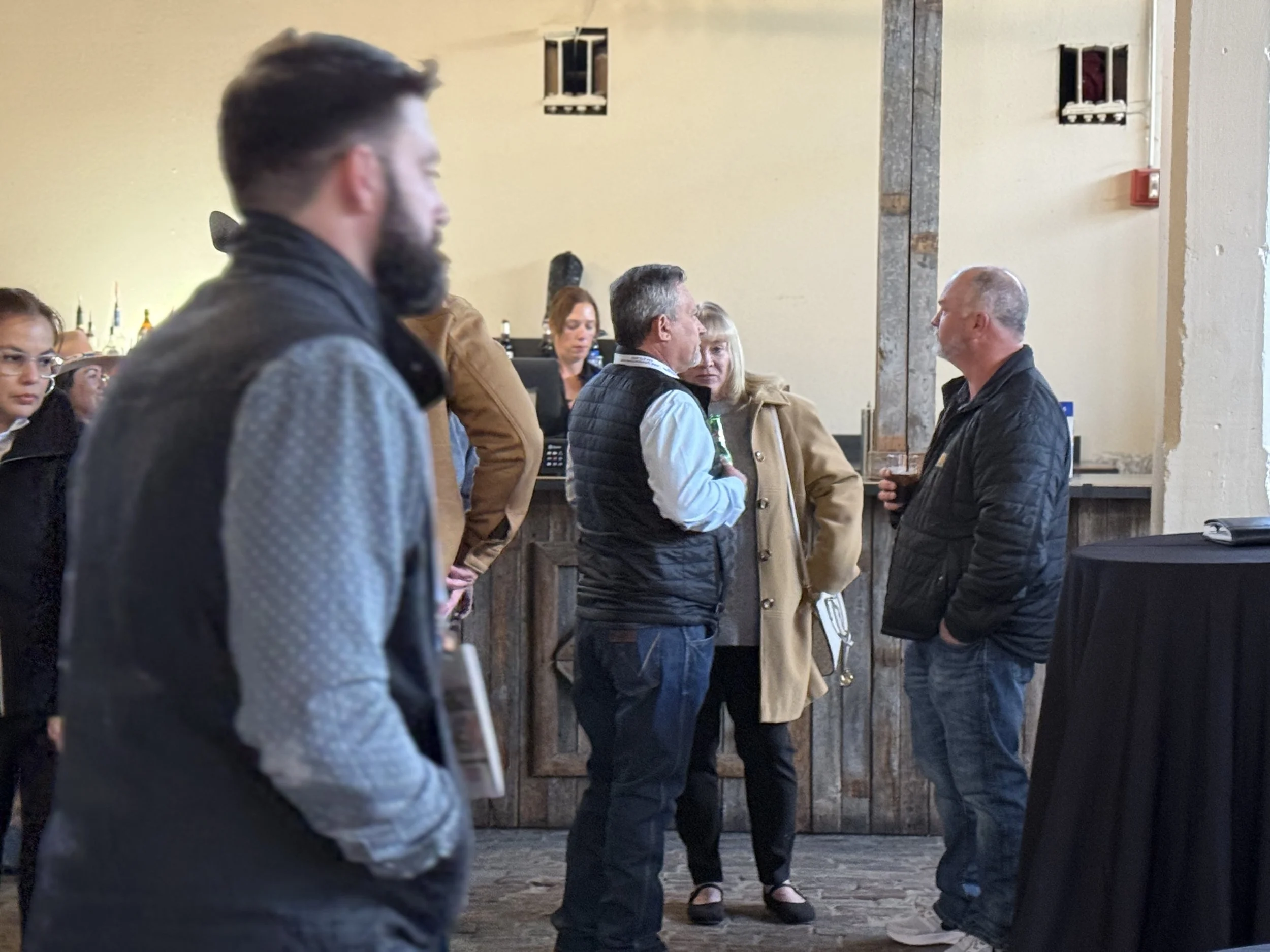 Several people are standing and talking in a room with a bar in the background. They are wearing casual and semi-casual clothing. The setting appears to be a social gathering or event.