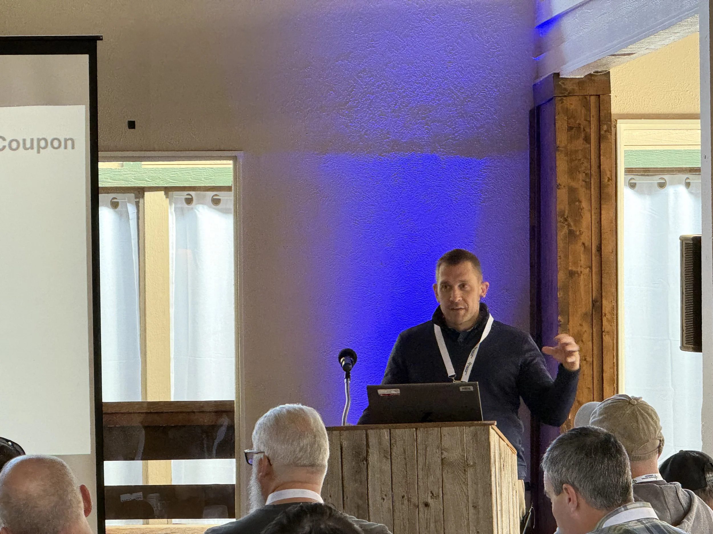 A man giving a presentation at a conference, standing behind a wooden podium with a laptop, in a room with large windows and an audience of diverse people.