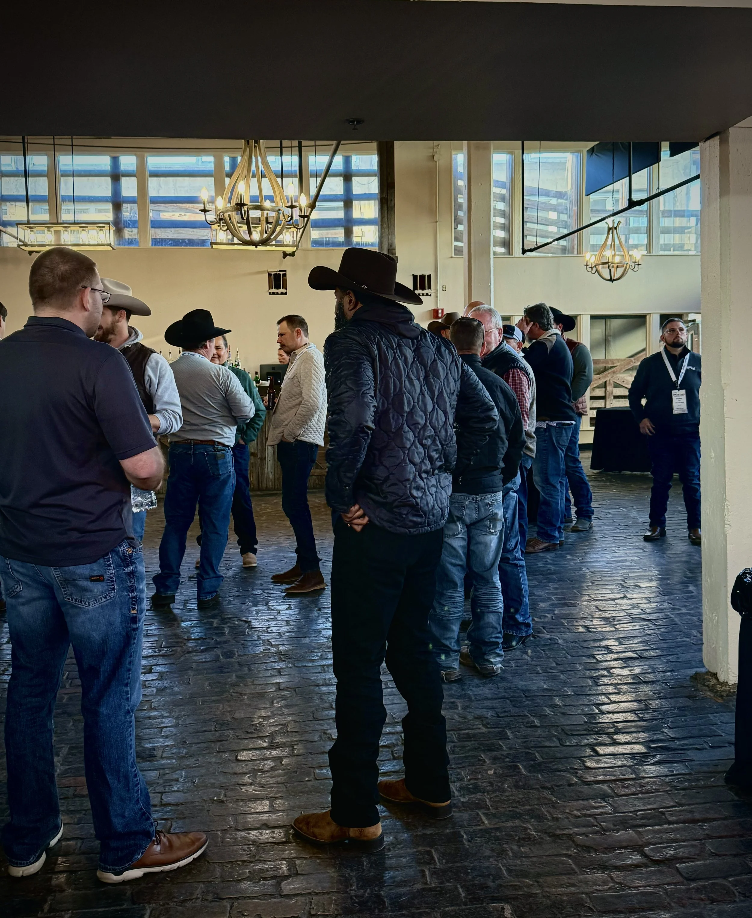 Group of people with cowboy hats and casual attire socializing indoors with large windows and chandeliers.