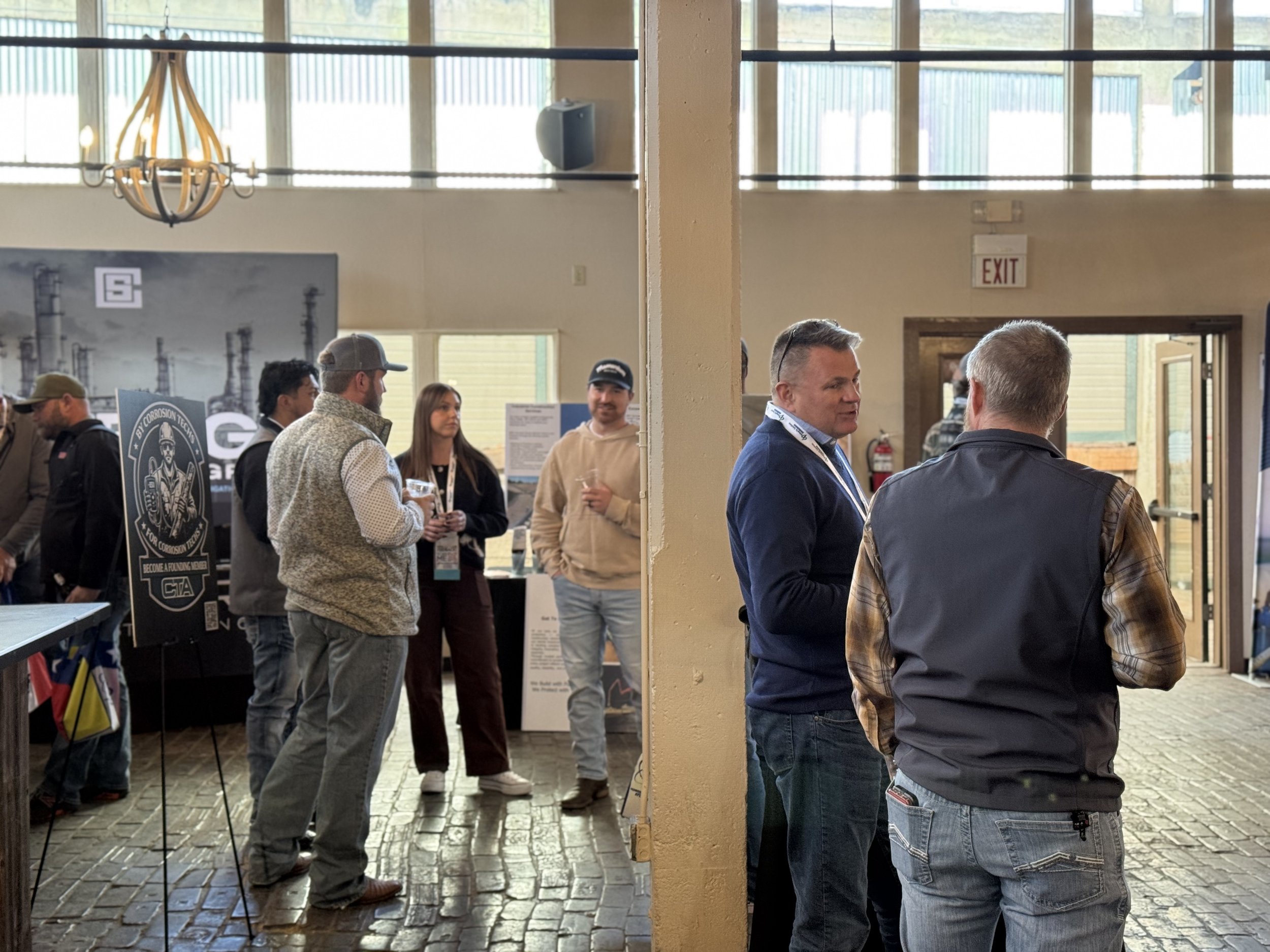 People attending an indoor event or exhibition, talking and socializing near display booths, with sunlight coming through large windows.