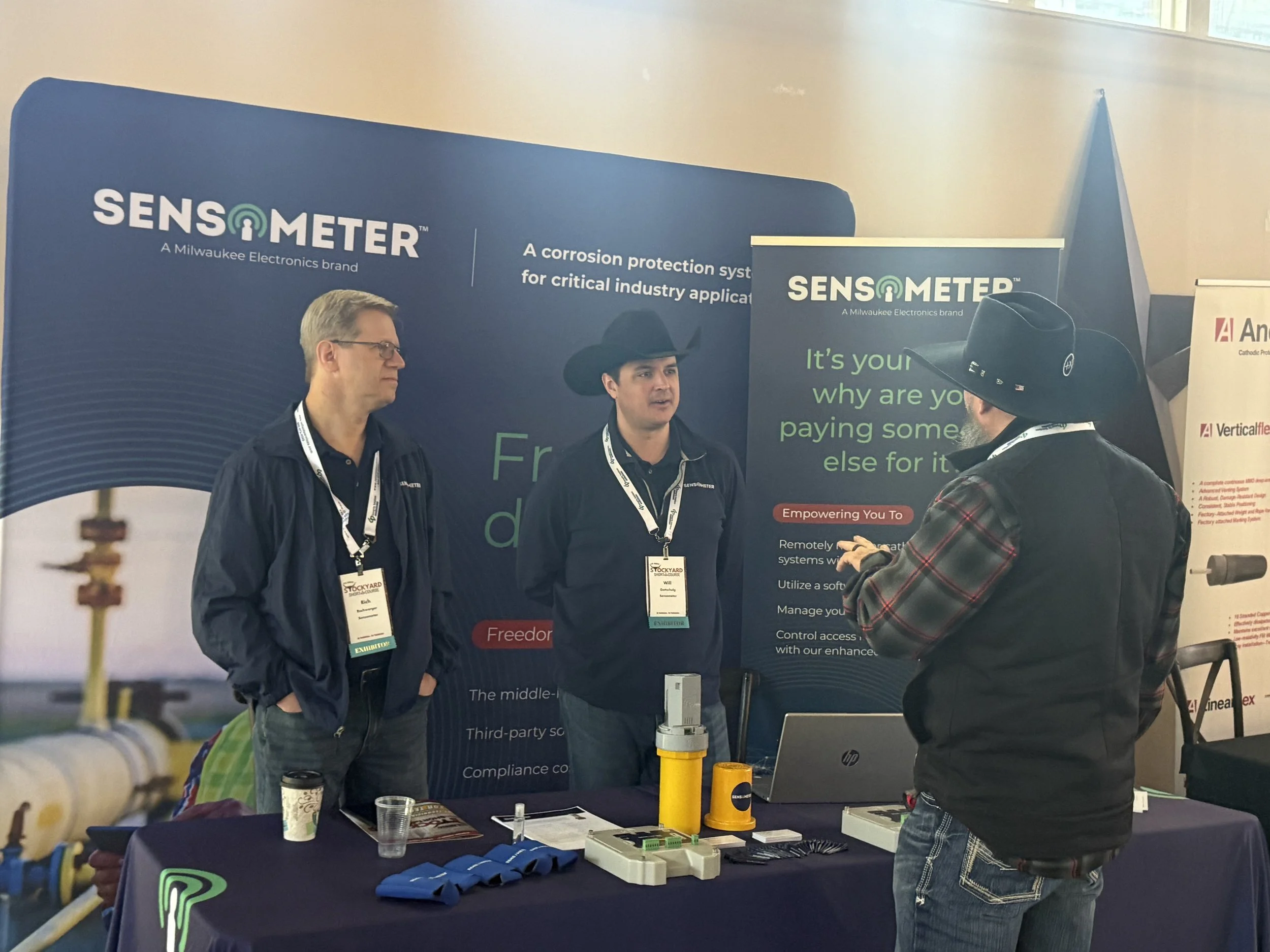 Three men at a trade show booth discussing industrial sensors, with banners displaying the SENSOMETER logo and product details.