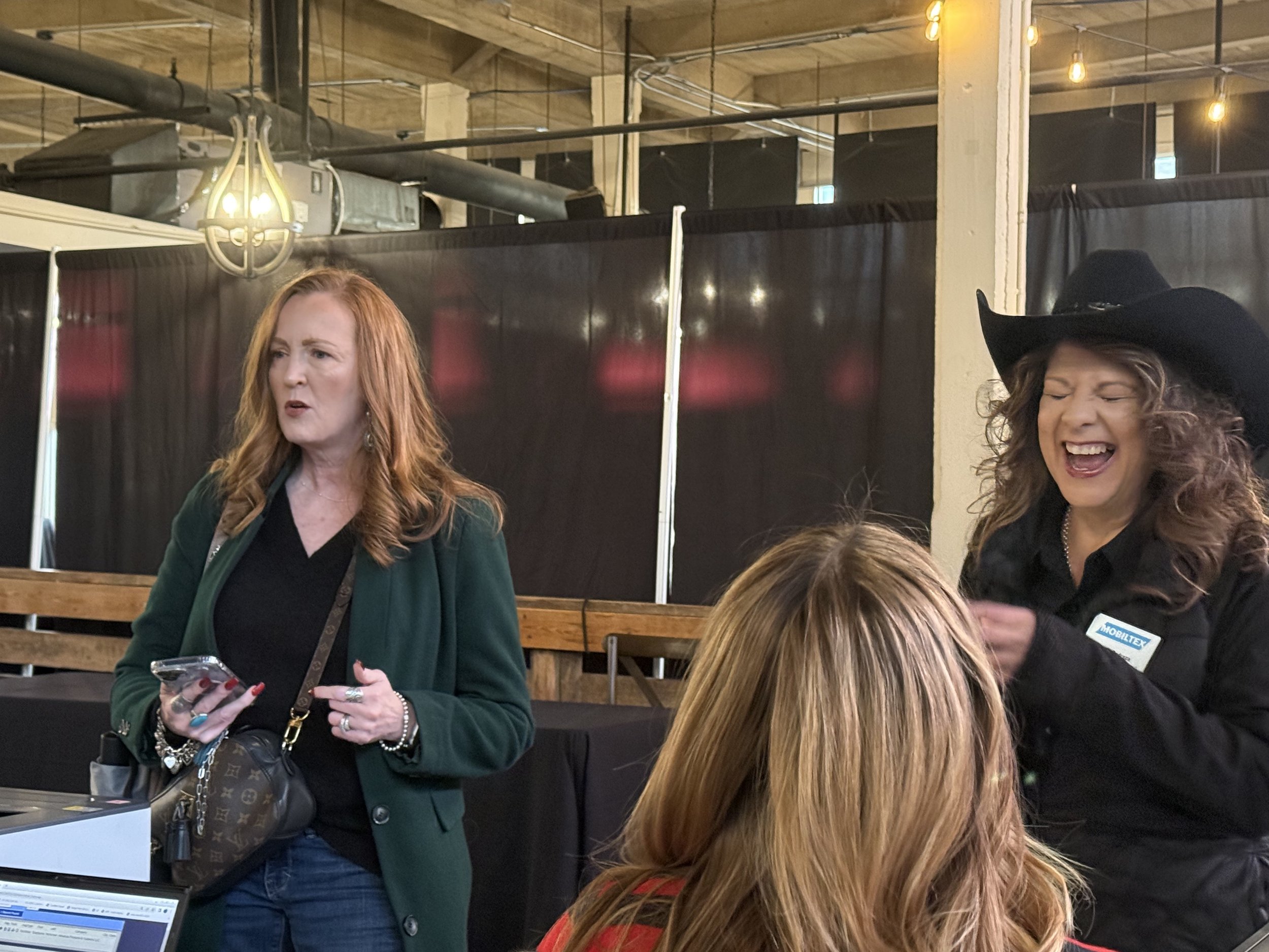 Two women at an indoor event, one with red hair in a green jacket holding a phone, the other with curly hair in a black cowboy hat laughing.