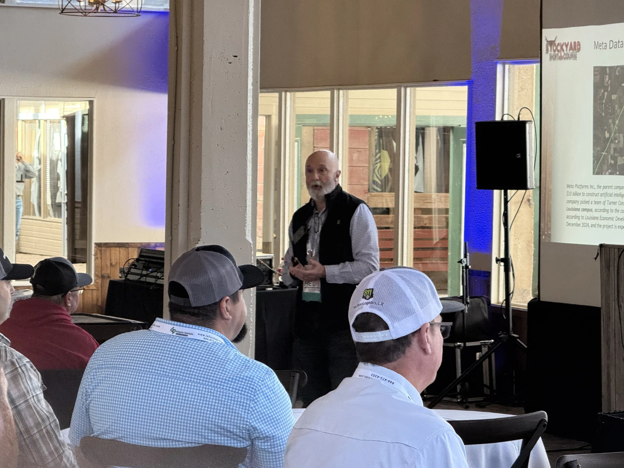 A man giving a presentation to an audience in a room with large windows, some audience members wearing hats, and a projection screen displaying a slide titled 'Meta Data'.