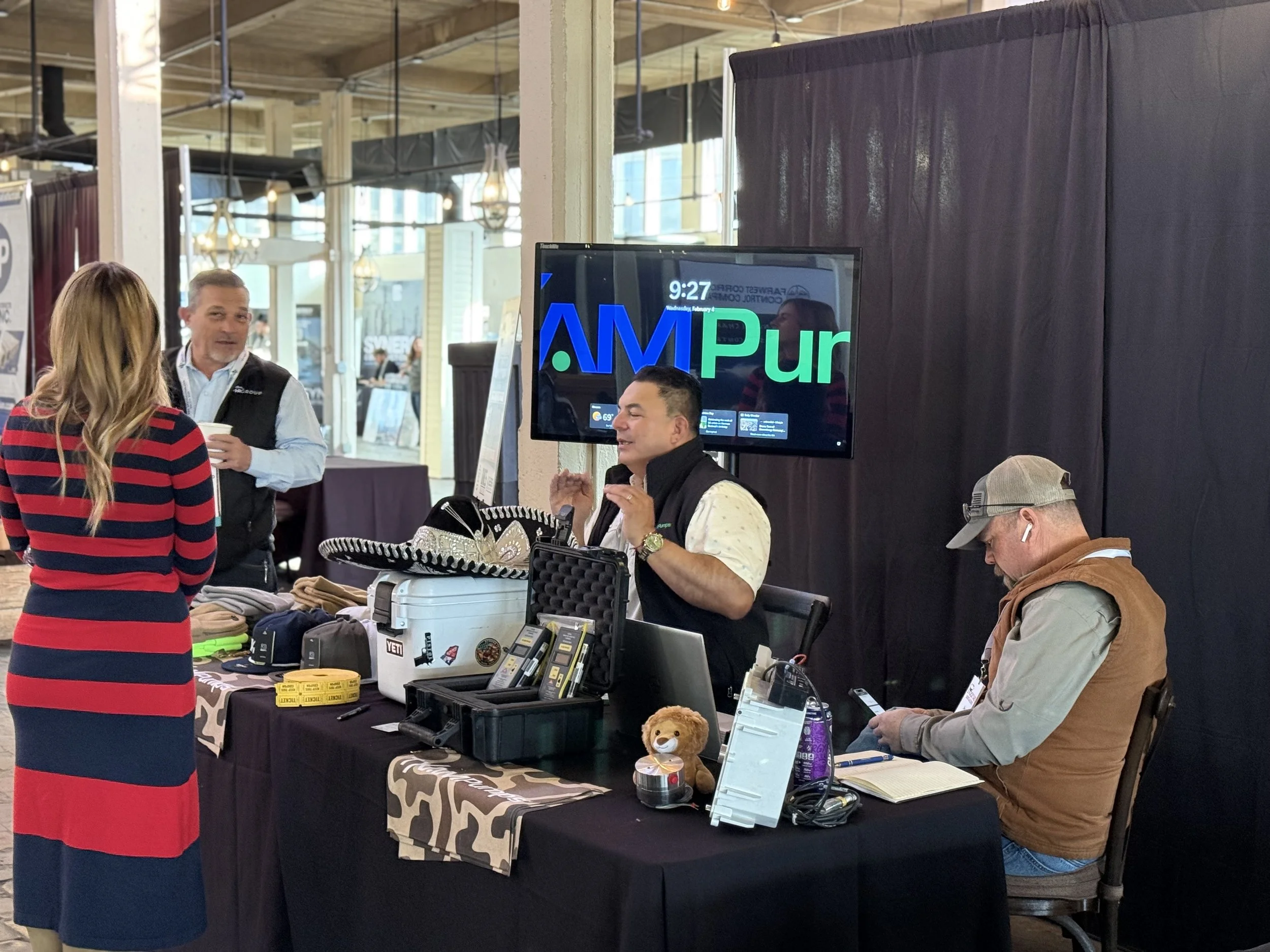 People at a booth with merchandise, a man in a vest and cowboy hat, a woman in a striped dress, and another man with a cap wearing AirPods, a flat-screen TV behind the booth displaying 'AM' in large blue and green letters, and various items on the ta