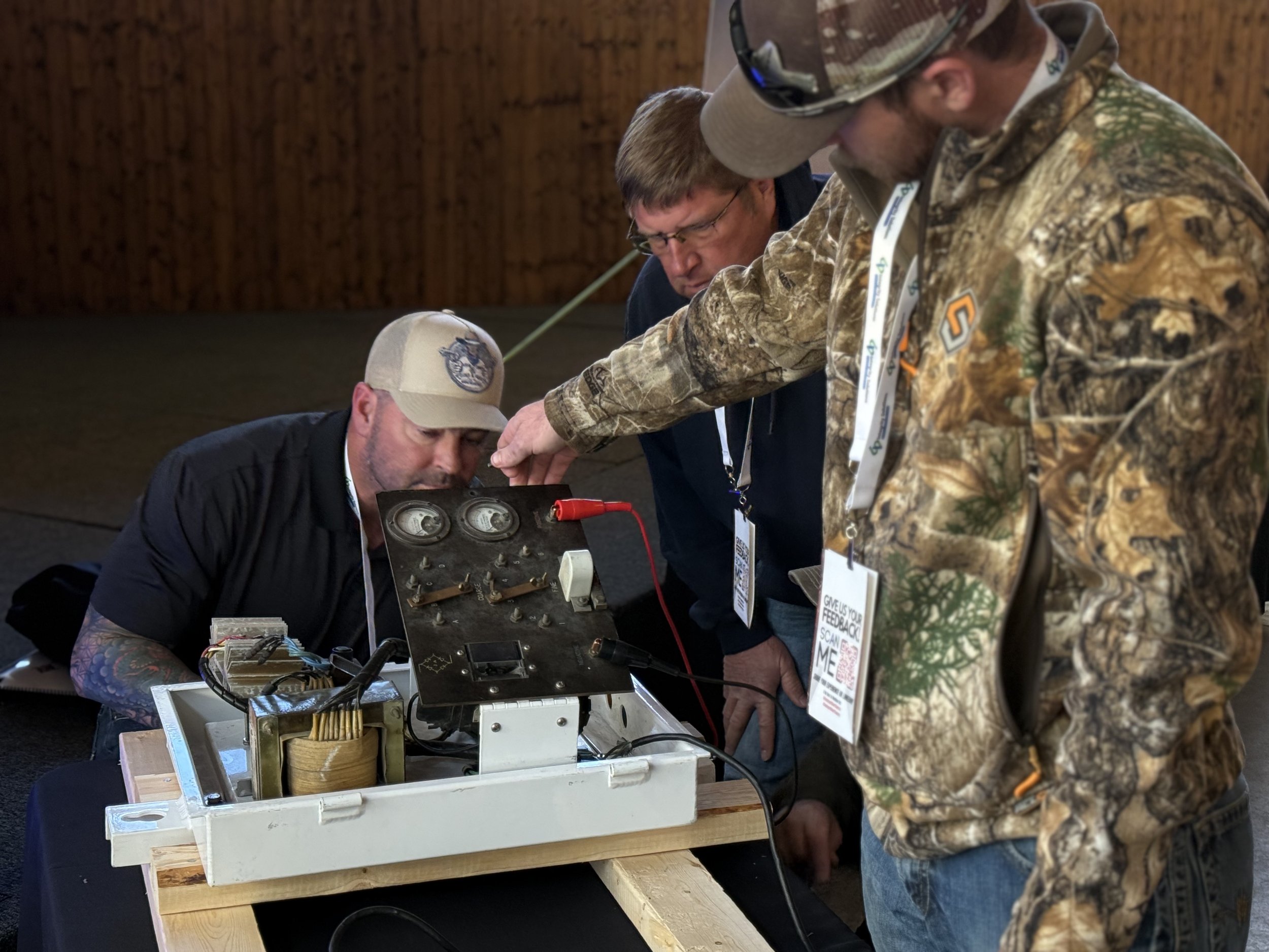 A group of four men working closely together on a technical device with wiring and gauges, with two men inspecting and working on the device and two men observing.