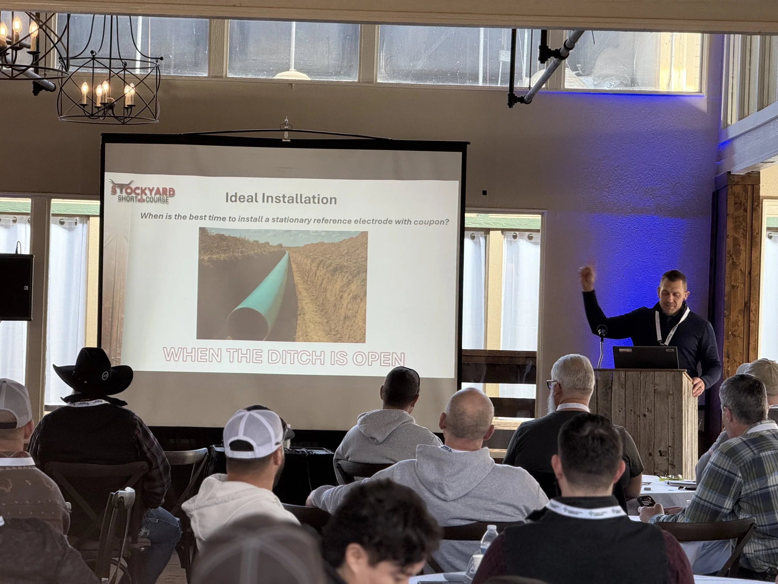 A man giving a presentation in a conference room with a projection screen displaying a slide titled "Ideal Installation". The slide shows a large pipe and the text "WHEN THE DITCH IS OPEN" at the bottom. The audience is seated and paying attention.