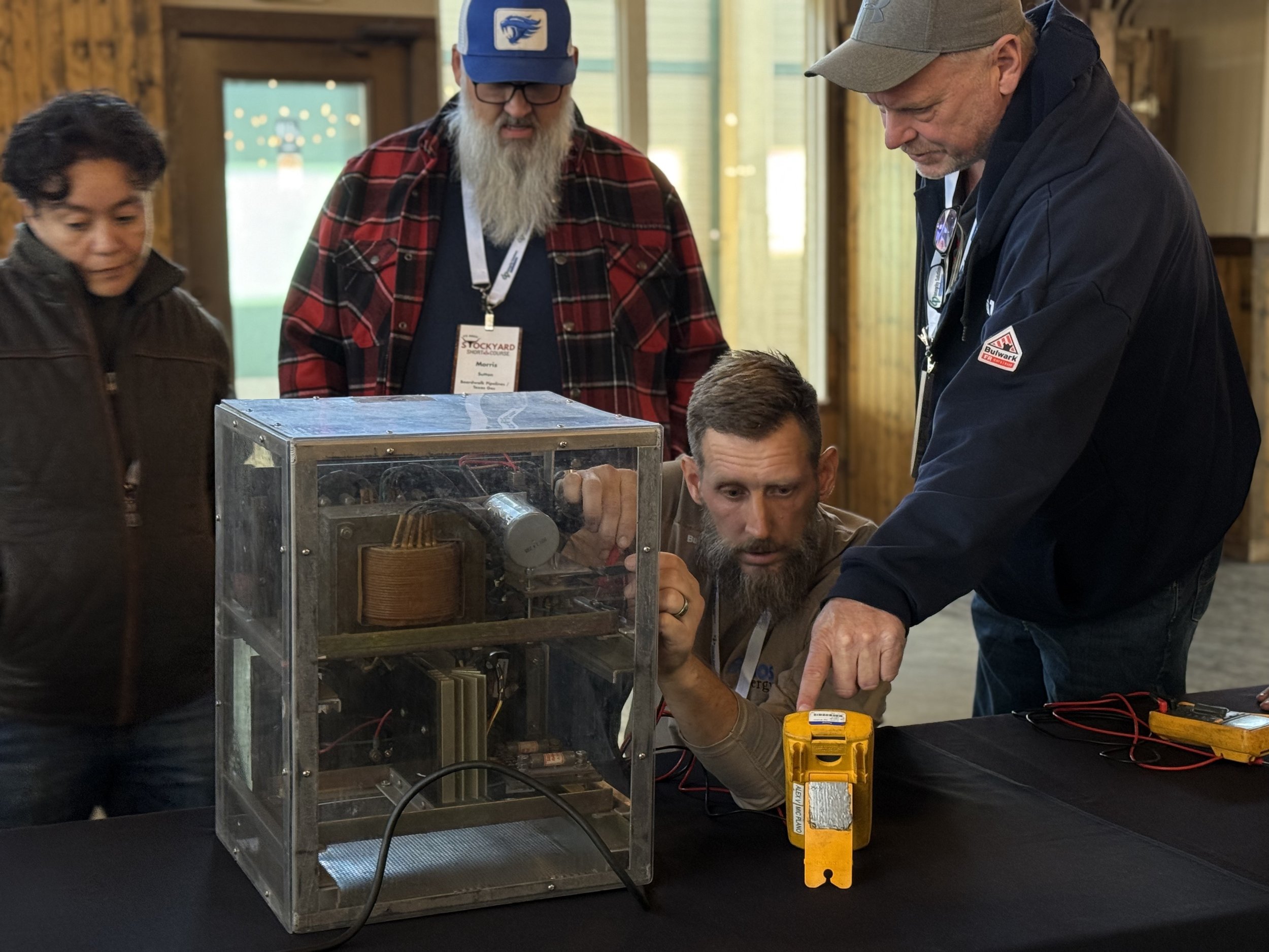 Group of five adults working on an electronics project with a transparent box on a black table, using a multimeter, in a rustic indoor setting.