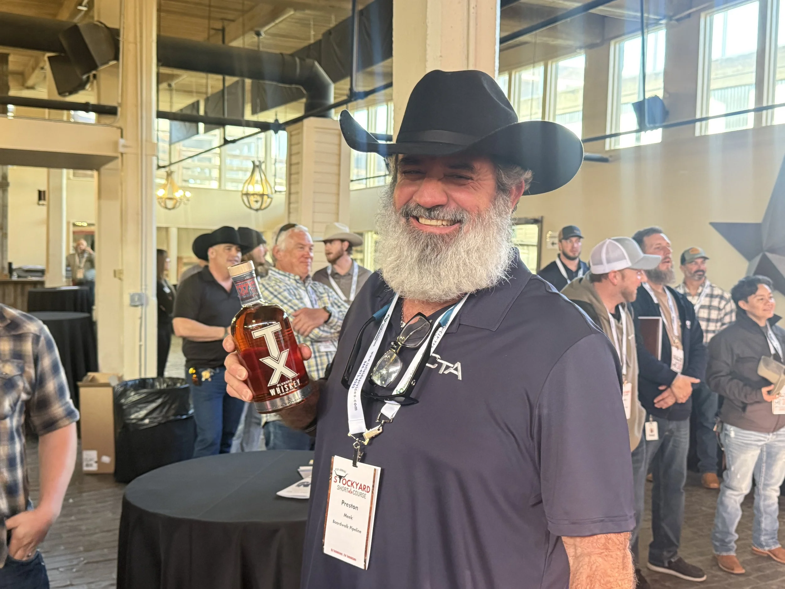A smiling man with a gray beard wearing a black cowboy hat and a navy blue polo shirt, holding a bottle of TX whiskey at an indoor event with other attendees in the background.