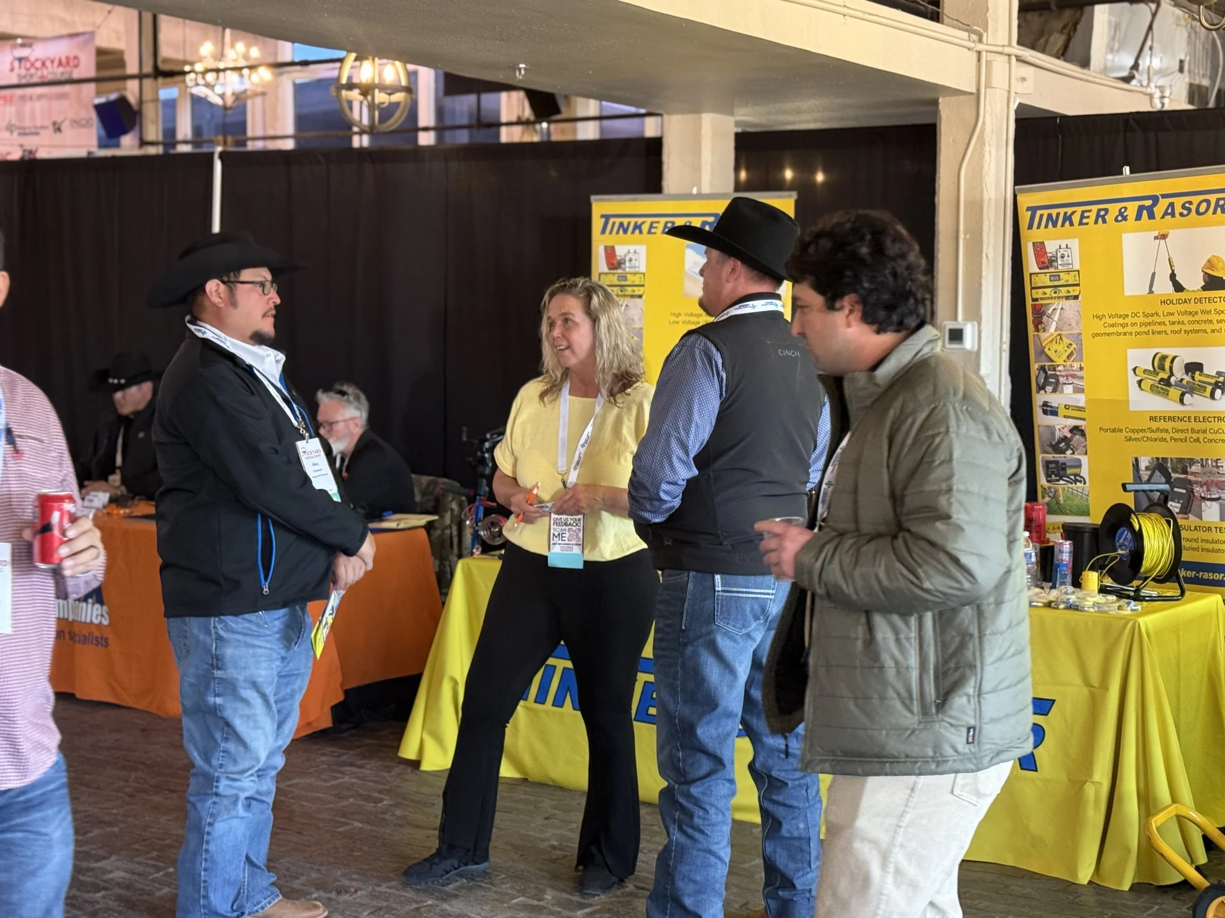 Three men and one woman are engaged in a conversation at an indoor trade show booth with yellow and black banners displaying Tinker & Rason products; the woman is smiling, the men are wearing casual jackets and hats.