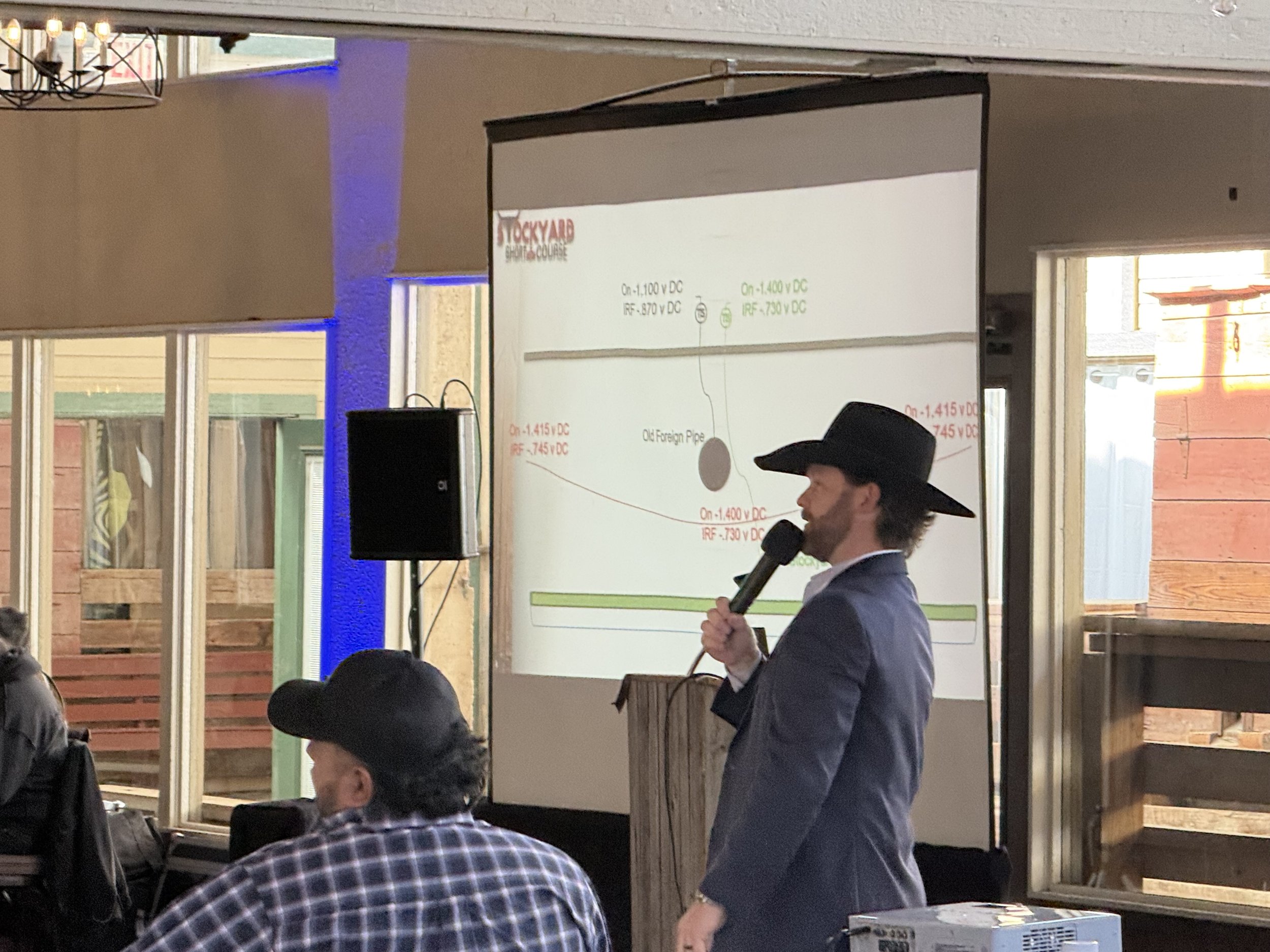 A man in a dark suit and black cowboy hat speaking into a microphone at a presentation. The presentation slide is projected on a screen behind him, showing technical information and diagrams. Several people are seated, one wearing a plaid shirt and a