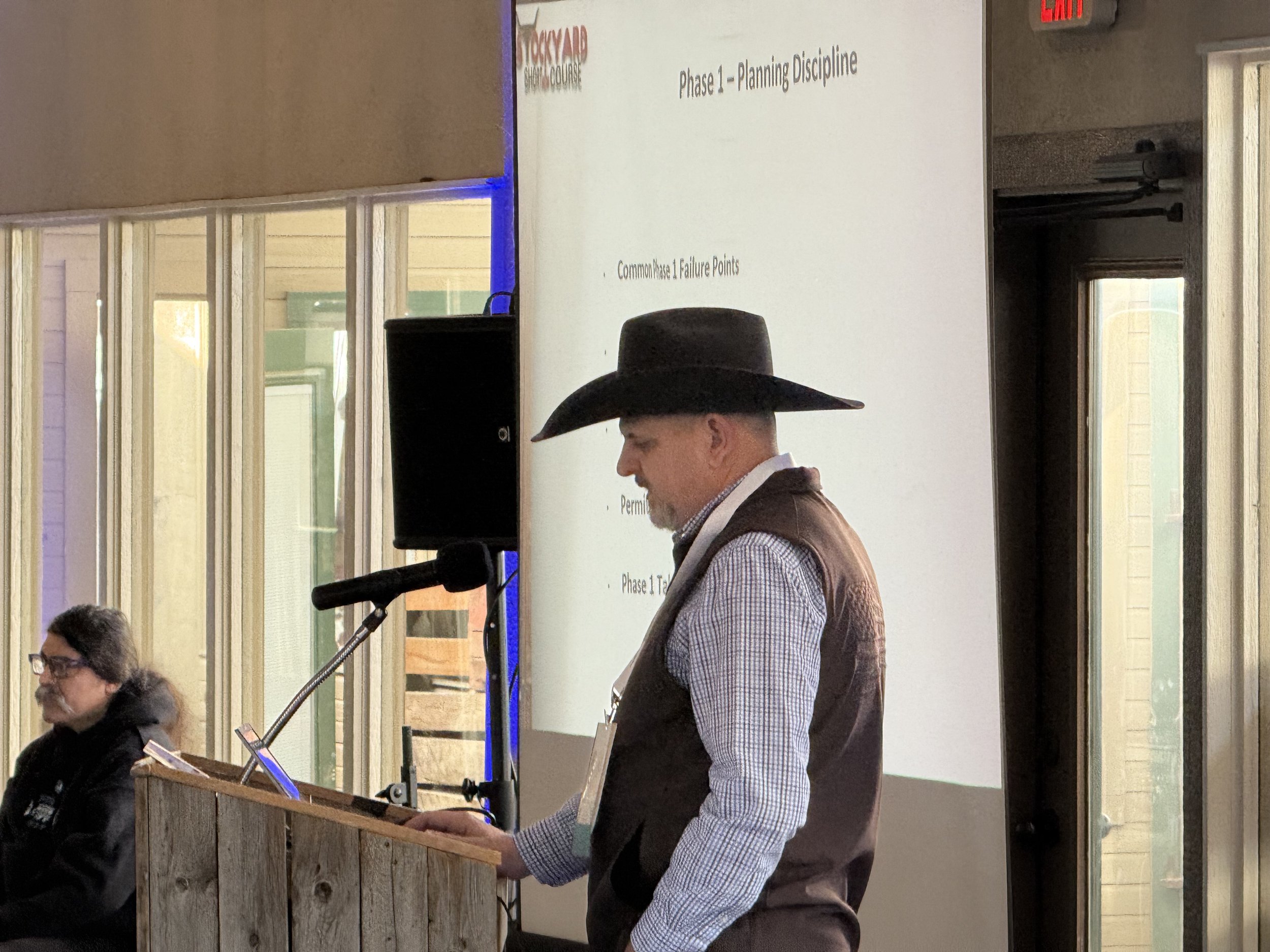 A man in a cowboy hat and checkered shirt standing next to a podium, reading from a paper at a presentation or event. In the background, there is a white screen with a presentation slide titled "Phase 1 - Planning Discipline." A woman with glasses an