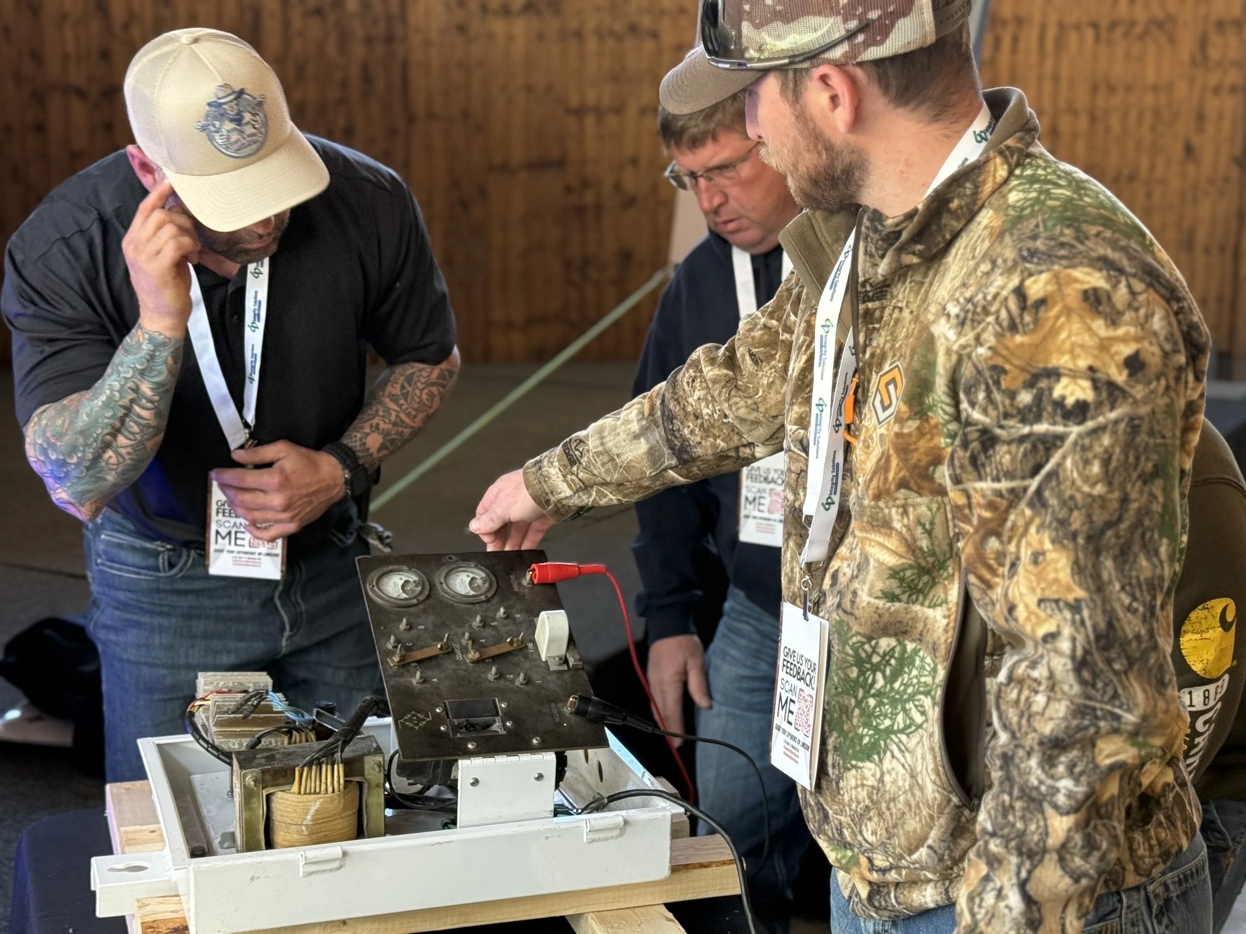 Three men observing and working with electronic equipment on a table at an indoor event. One man is in camouflage, another in dark clothing, and the third with tattoos and a baseball cap.