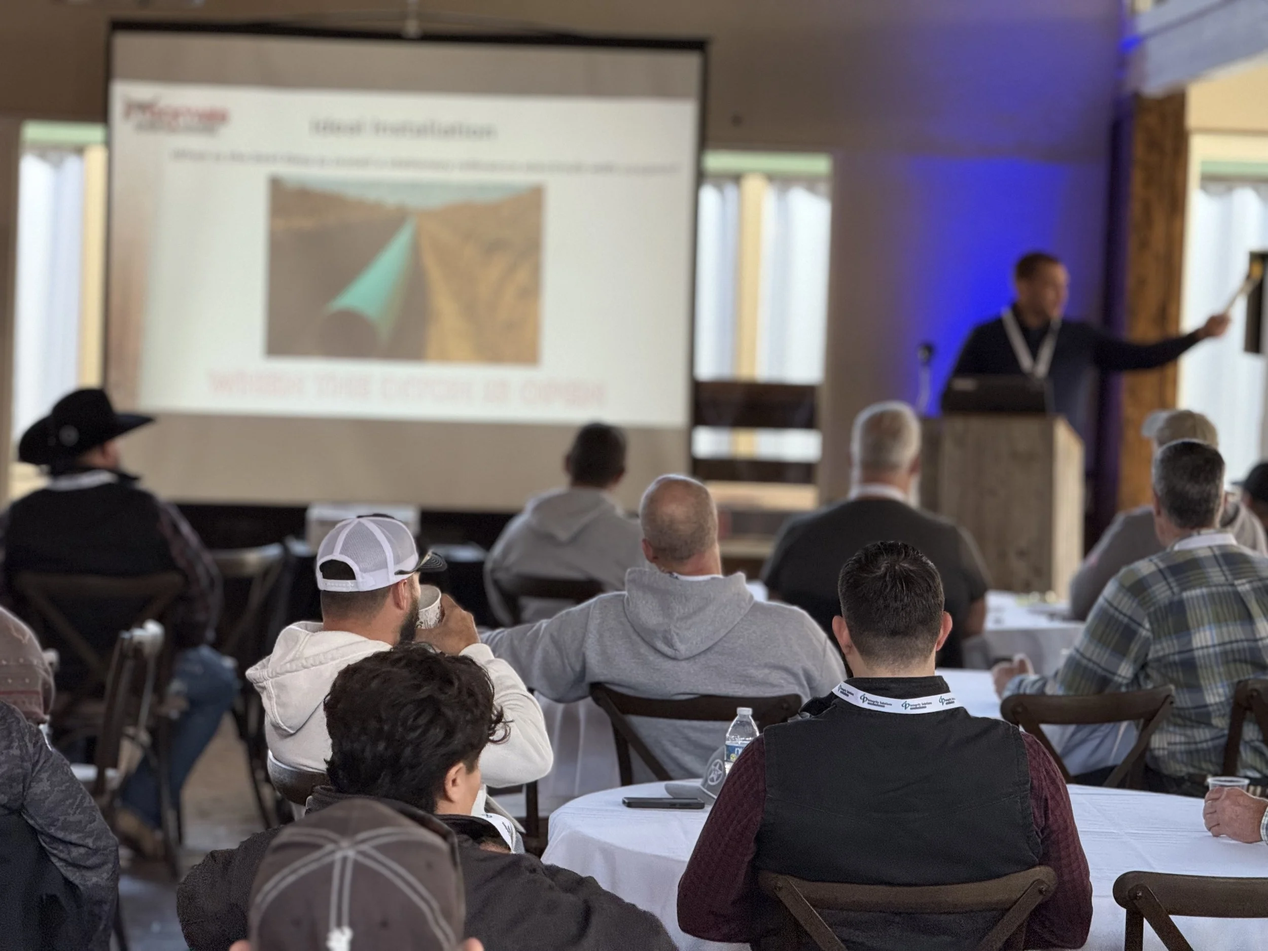 People seated at round tables in a conference room, watching a presentation. A presenter stands at a podium pointing at a screen displaying a slide with an image of a pipe in a desert landscape.