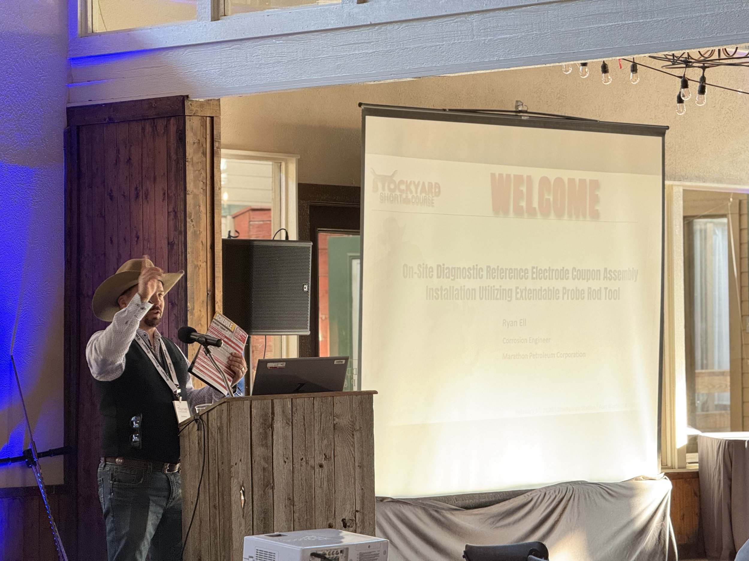 A man dressed as a cowboy, holding a brochure and speaking at a conference with a presentation screen behind him.