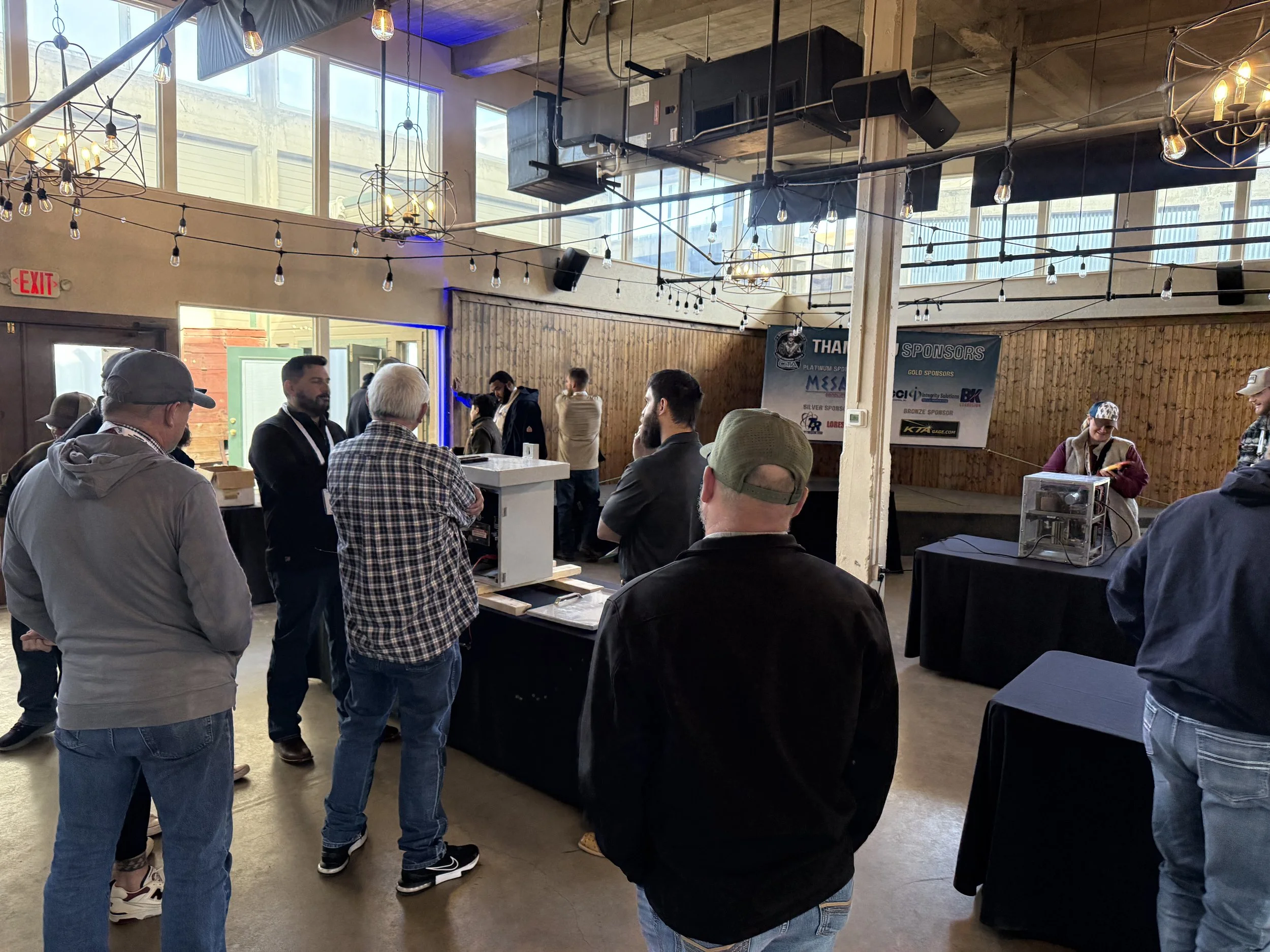 People standing in a line at an indoor event with string lights and a wooden wall. Some are engaged in conversation. There is a banner on the wall thanking sponsors, and a woman at a table with a display case.