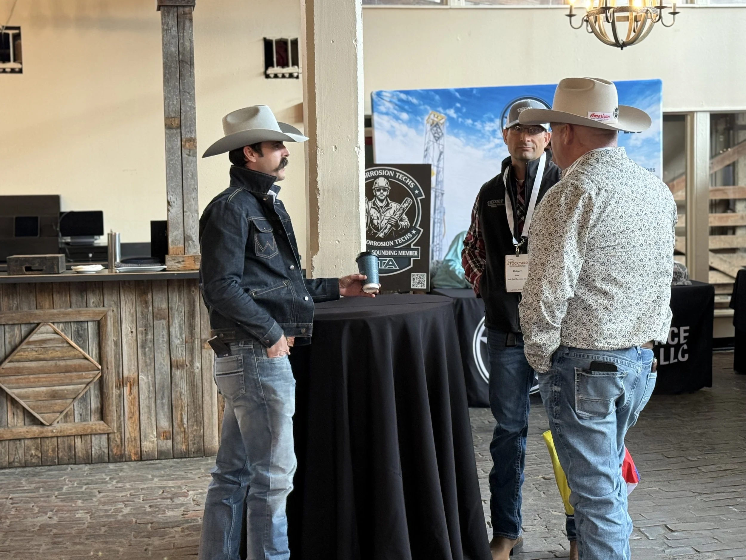 Three men wearing cowboy hats and casual clothes are having a conversation at an indoor event. One man is holding a cup, and there are posters and a bar in the background.