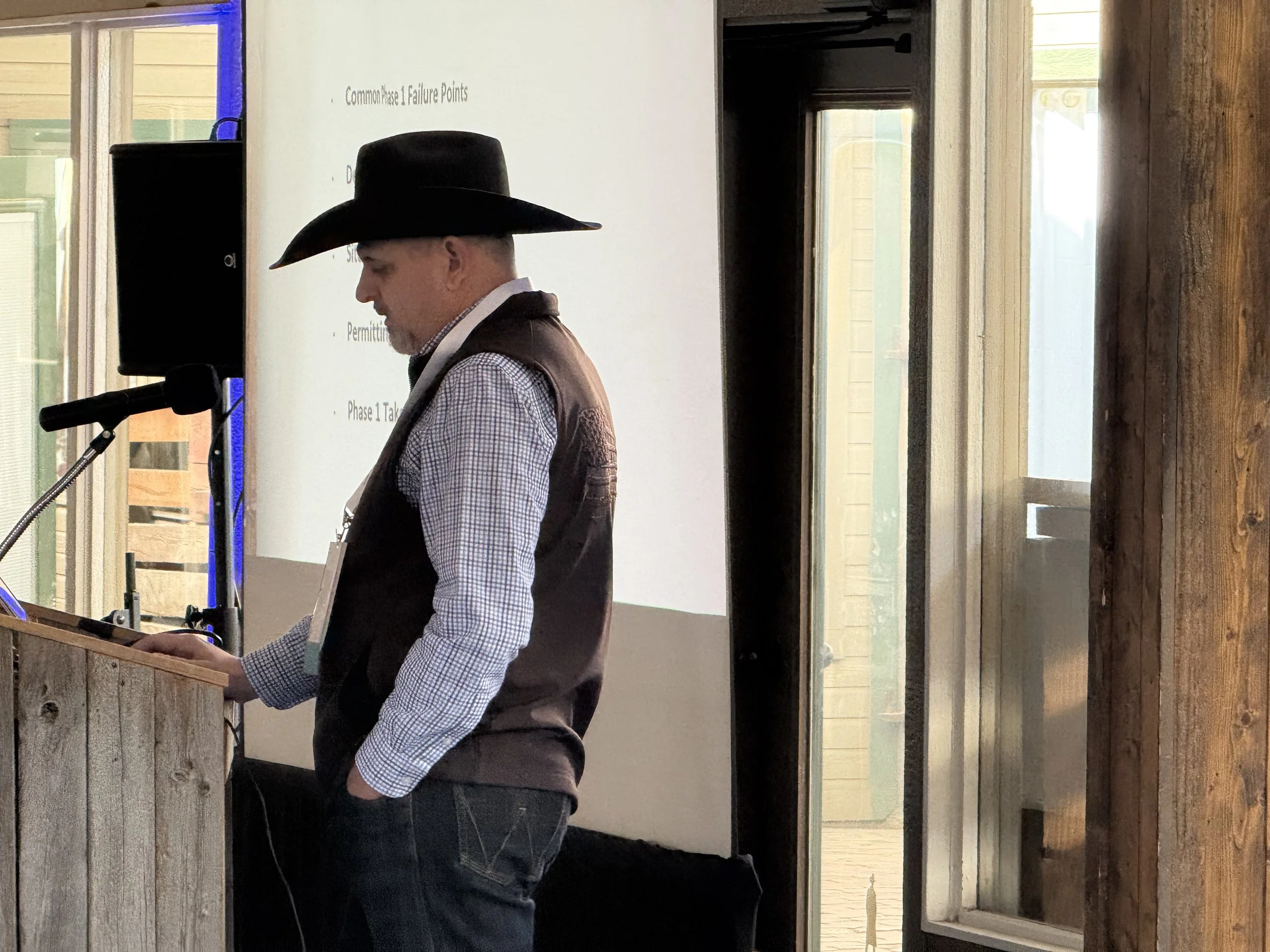 A man wearing a cowboy hat and checkered shirt giving a presentation in front of a projected slide that reads 'Common Phase 1 Failure Points' in a rustic wooden interior.
