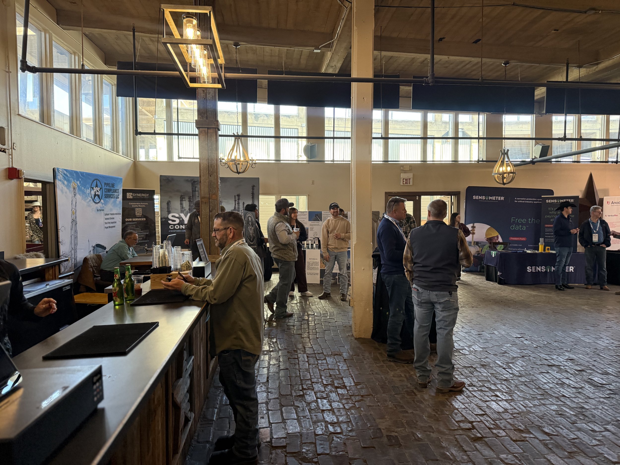 Indoor conference or trade show with booths and attendees talking, some using smartphones, in a brick-floored room with high ceilings and large windows.