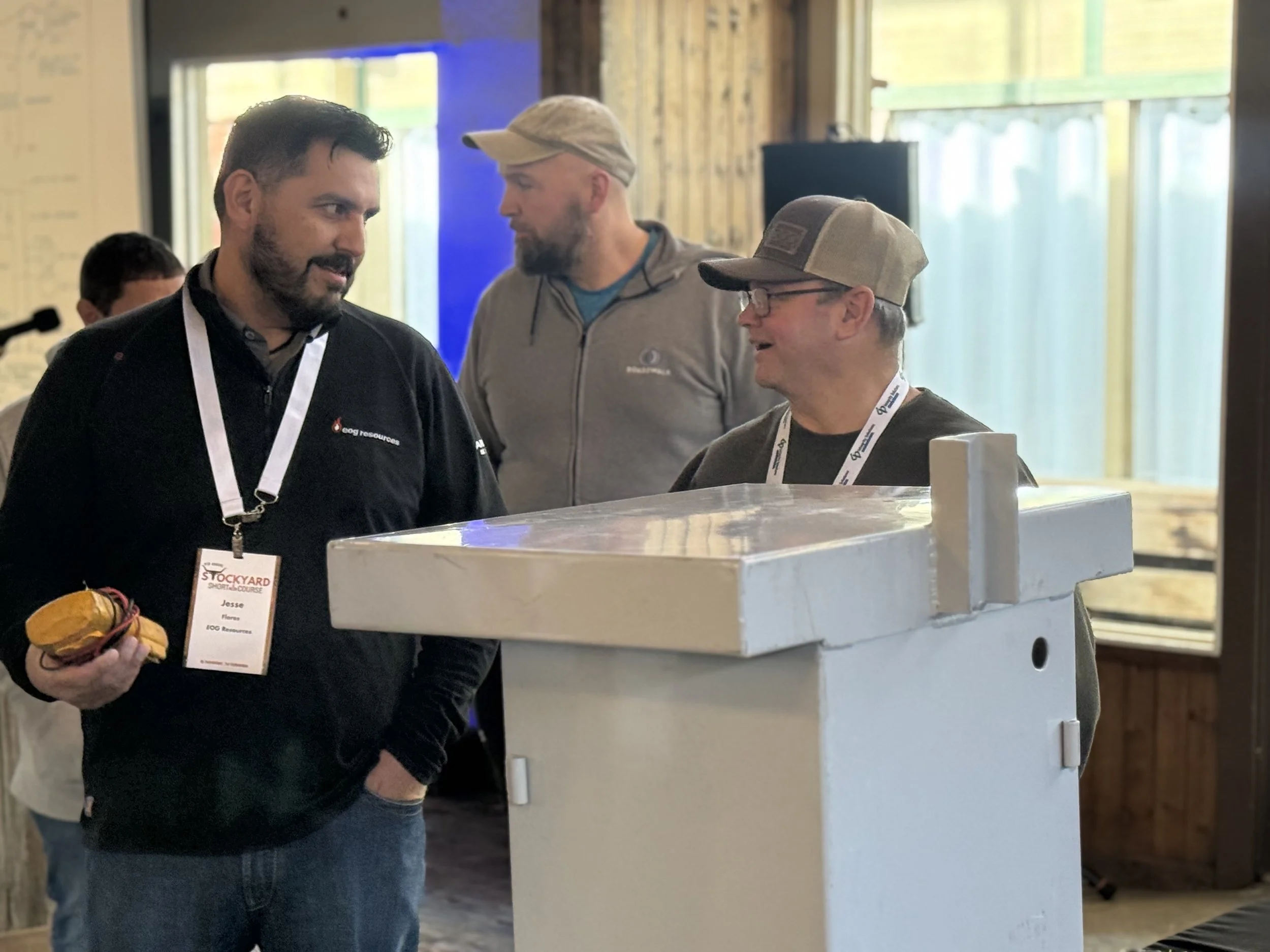 Three men conversing at an indoor event, one holding a yellow device, with a Rusty the Scarecrow sculpture in the background.