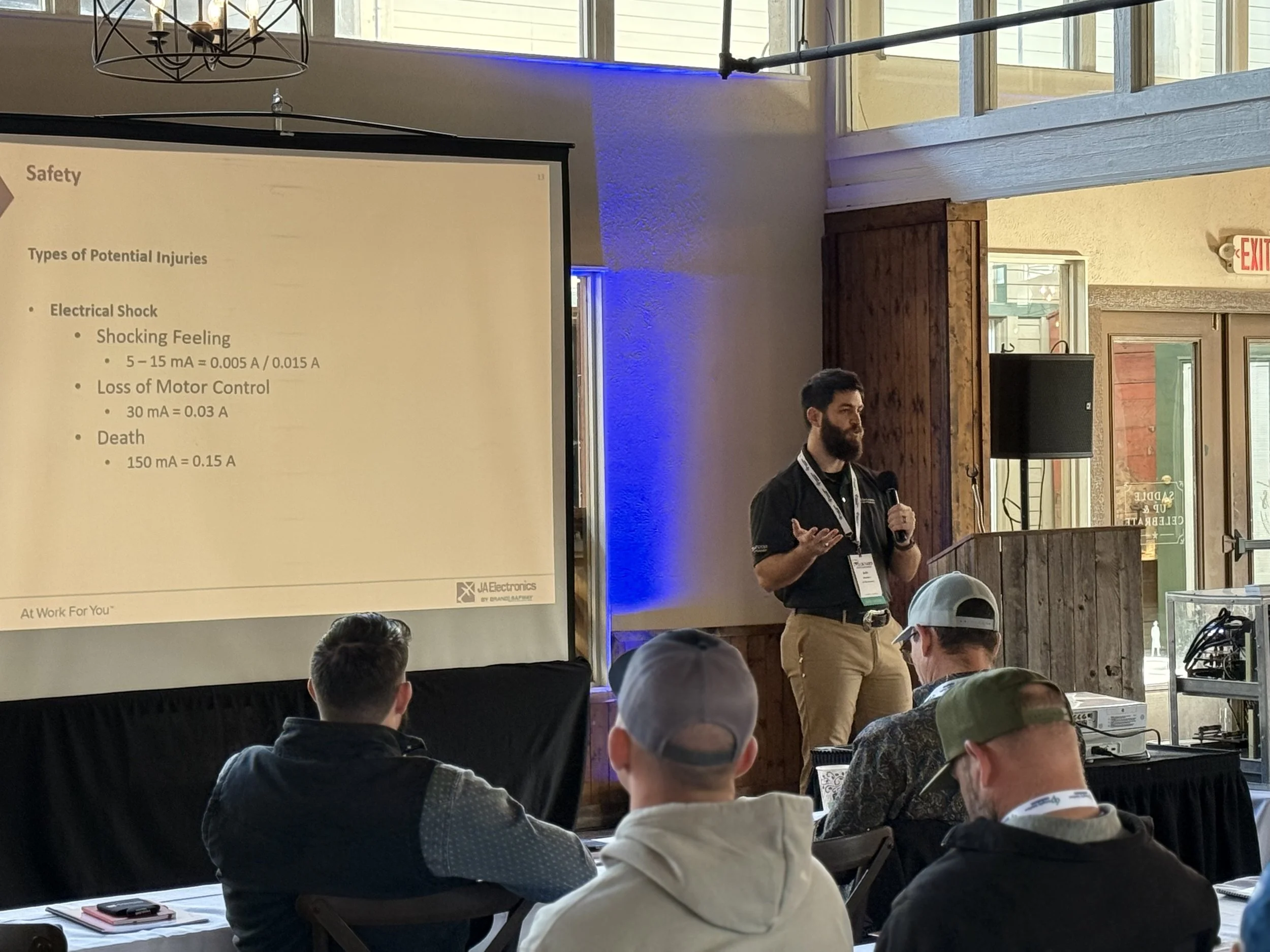 A man giving a presentation in a conference room with a PowerPoint slide about potential injuries from electrical shock. The room has natural light, wood accents, and an audience of people seated at tables.