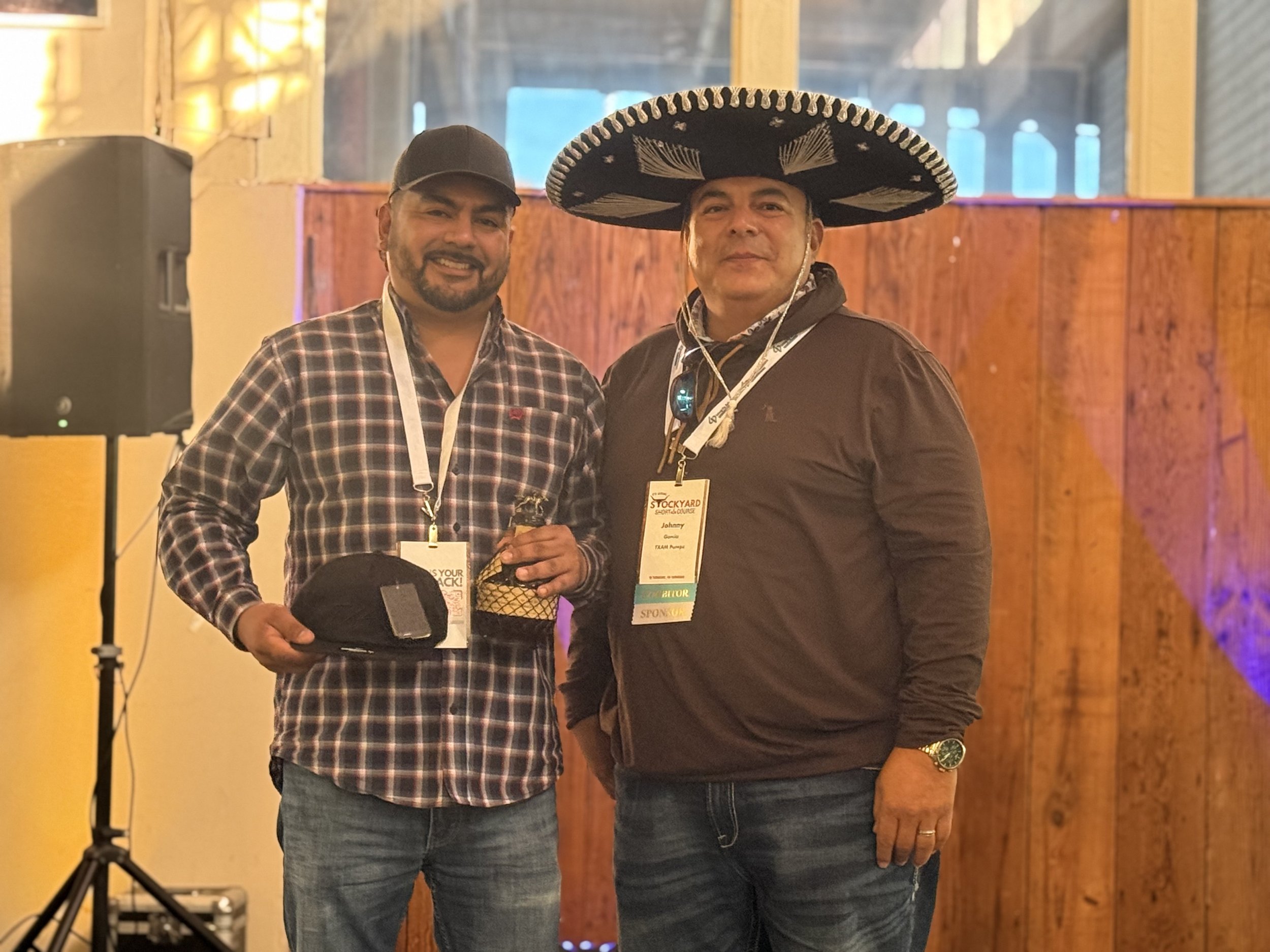 Two men standing side by side smiling at a conference, one wearing a black sombrero and the other holding a black cap, in front of a wooden wall.
