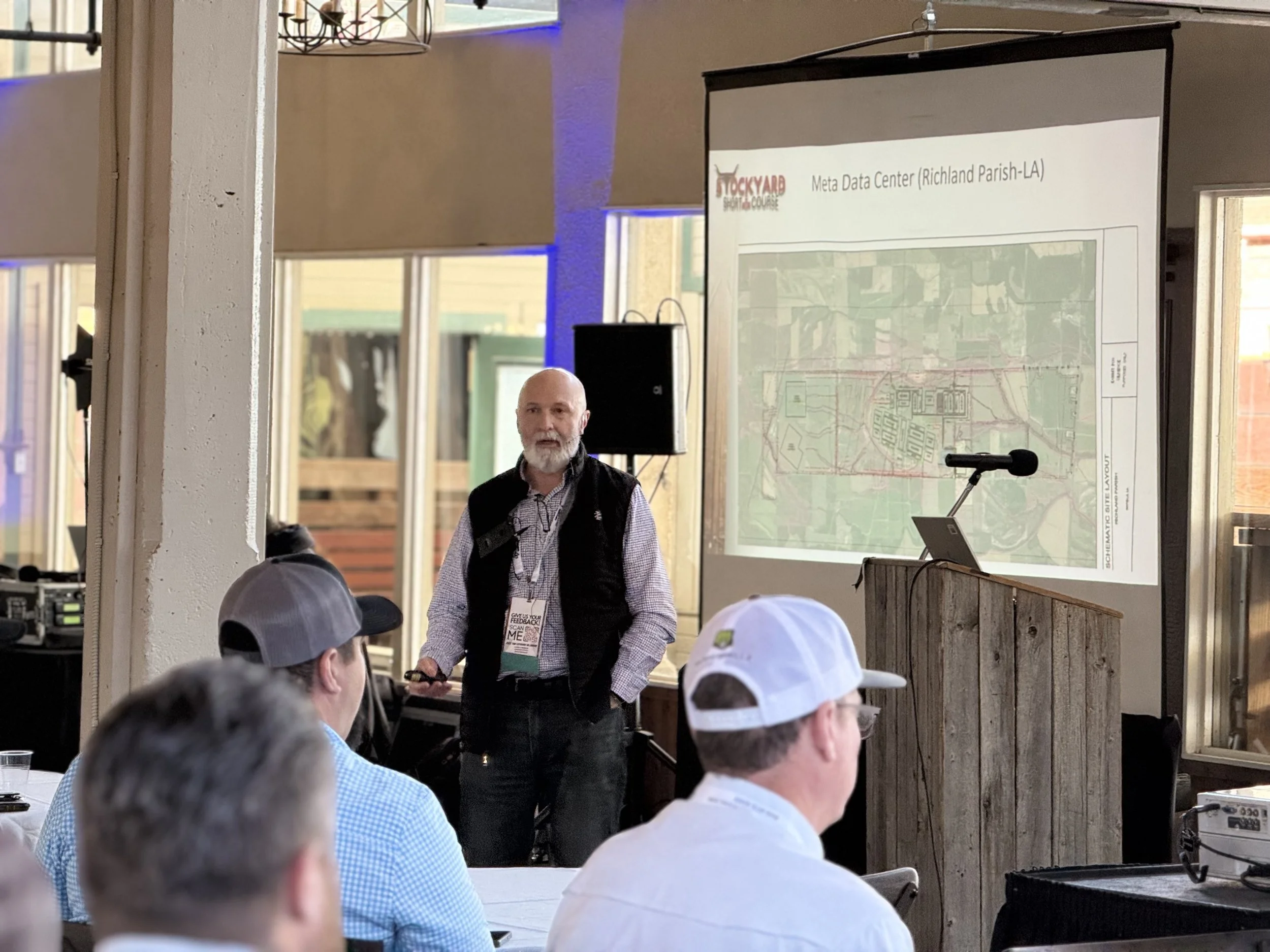 A man giving a presentation at a conference, standing in front of a screen displaying a map labeled "Meta Data Center (Richland Parish-LA)." Several audience members watch and listen.