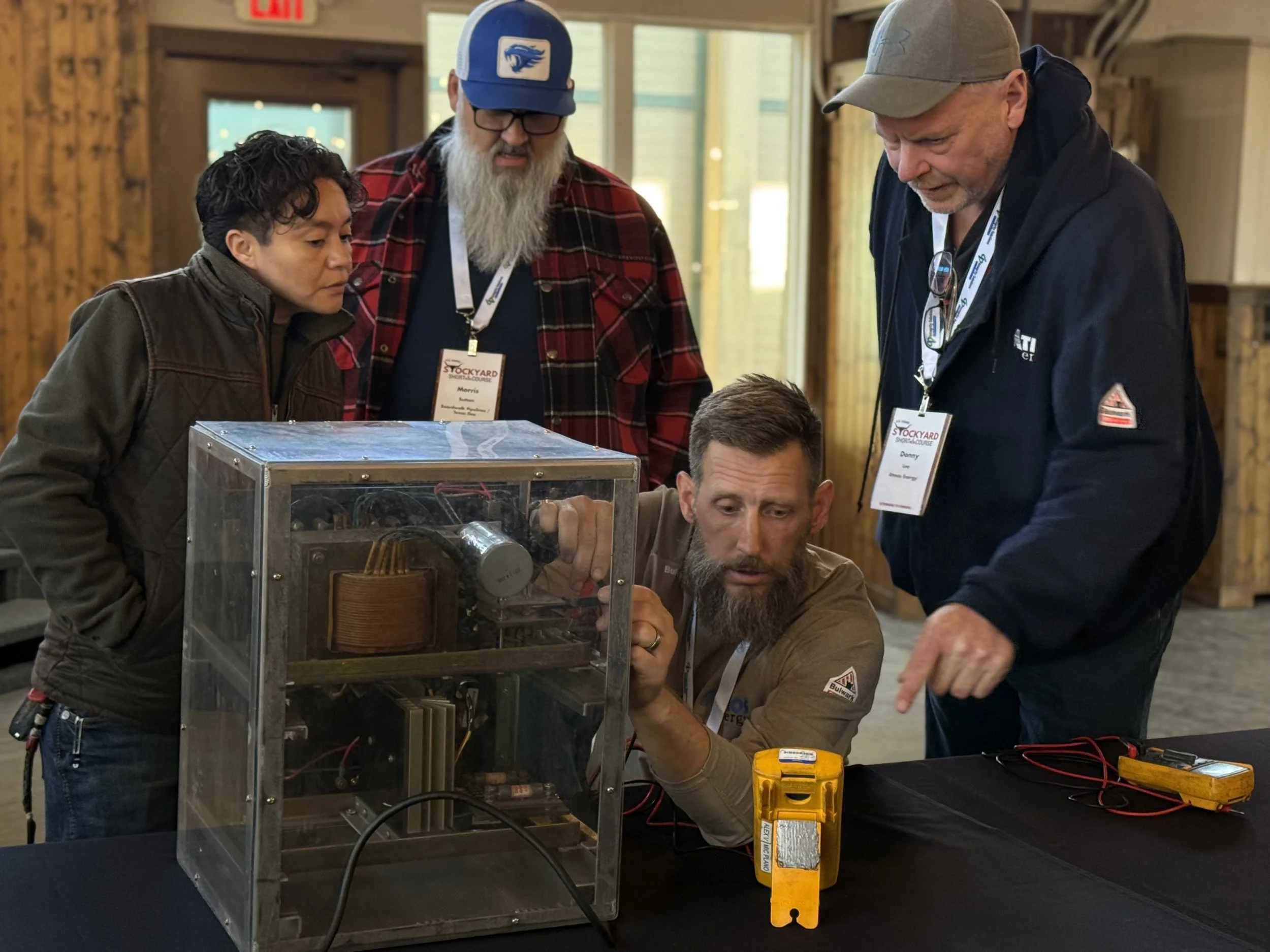 Four people gathered around a small, enclosed mechanical device during a demonstration. One man is seated and adjusting the device while three others observe closely. The scene appears to be in a rustic, indoor setting with wooden walls.