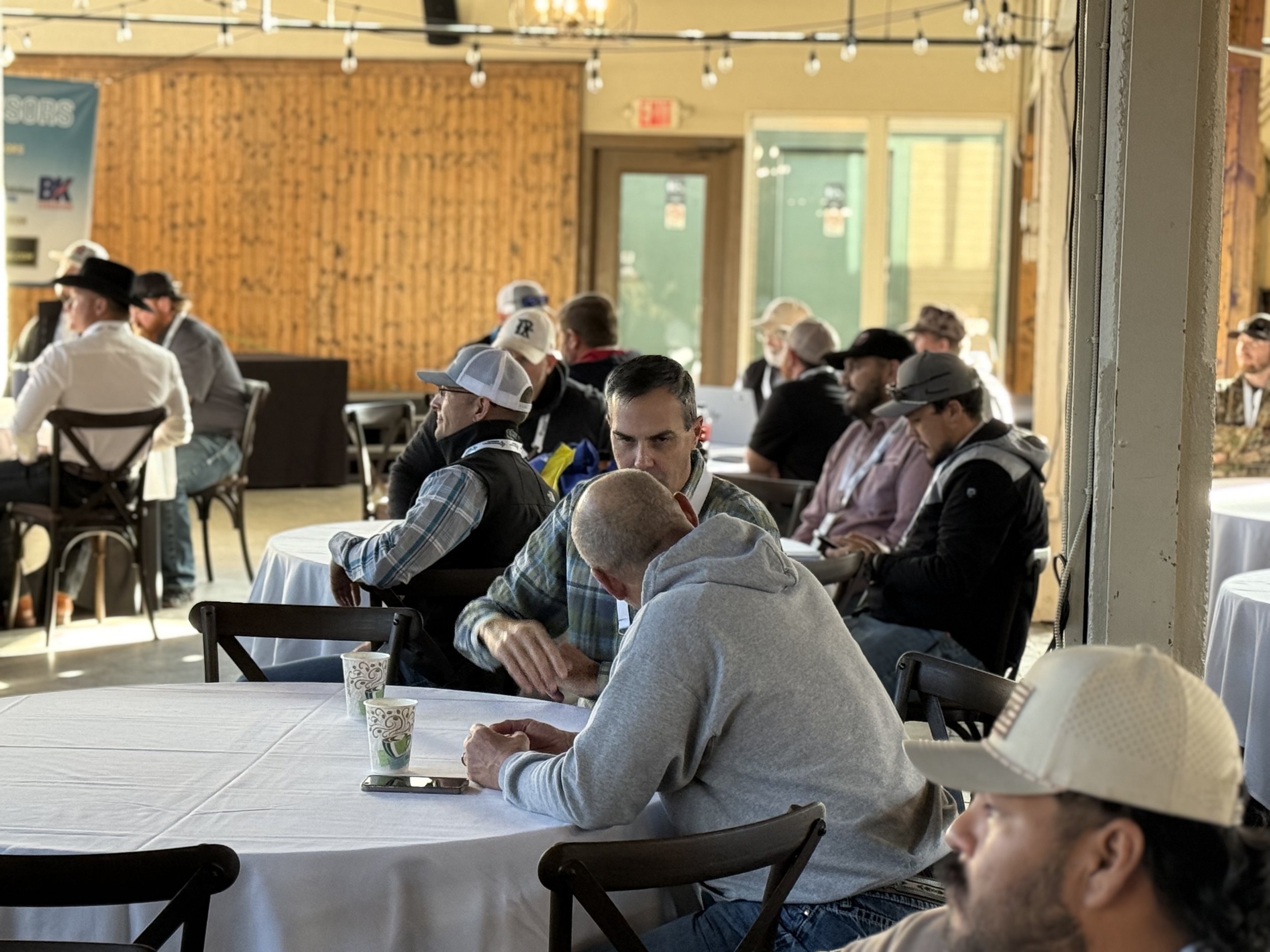 People sitting at round tables with white tablecloths in a wooden interior, some wearing hats and casual clothes, engaged in conversation or looking at their phones.