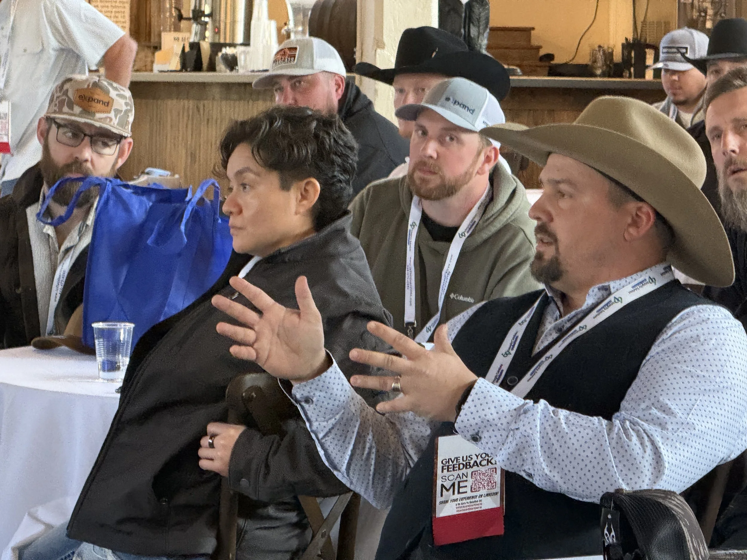 A group of people sitting at a table, engaged in a discussion or listening to a speaker. Some are wearing hats, and one man in the foreground is gesturing with his hands while wearing a wide-brimmed hat and a badge that reads 'Give us your feedback s