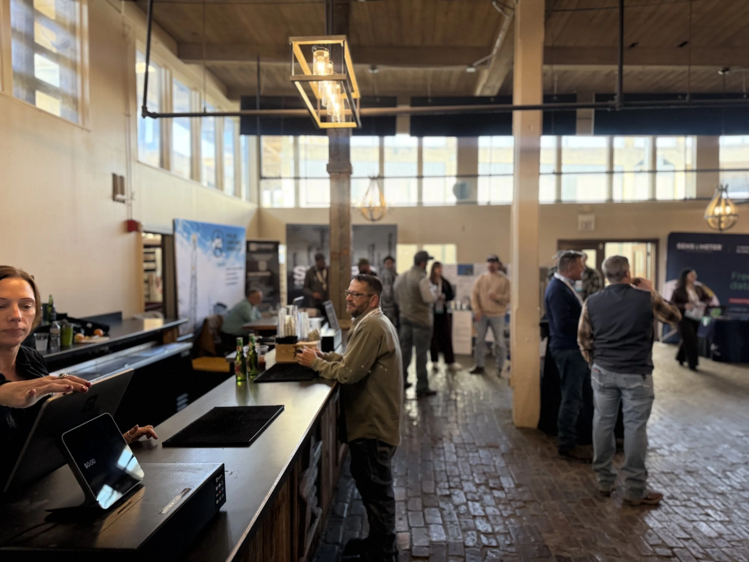 People gathered at a coffee bar inside a modern industrial-style building with high ceilings, large windows, and hanging lights.