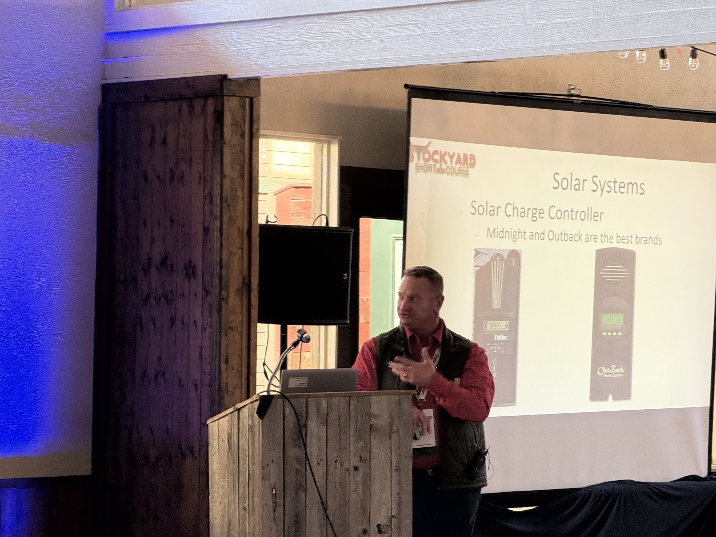 A man giving a presentation on solar systems at a conference, standing behind a wooden podium with a laptop, while a slide on the screen behind him discusses solar charge controllers with brand images.