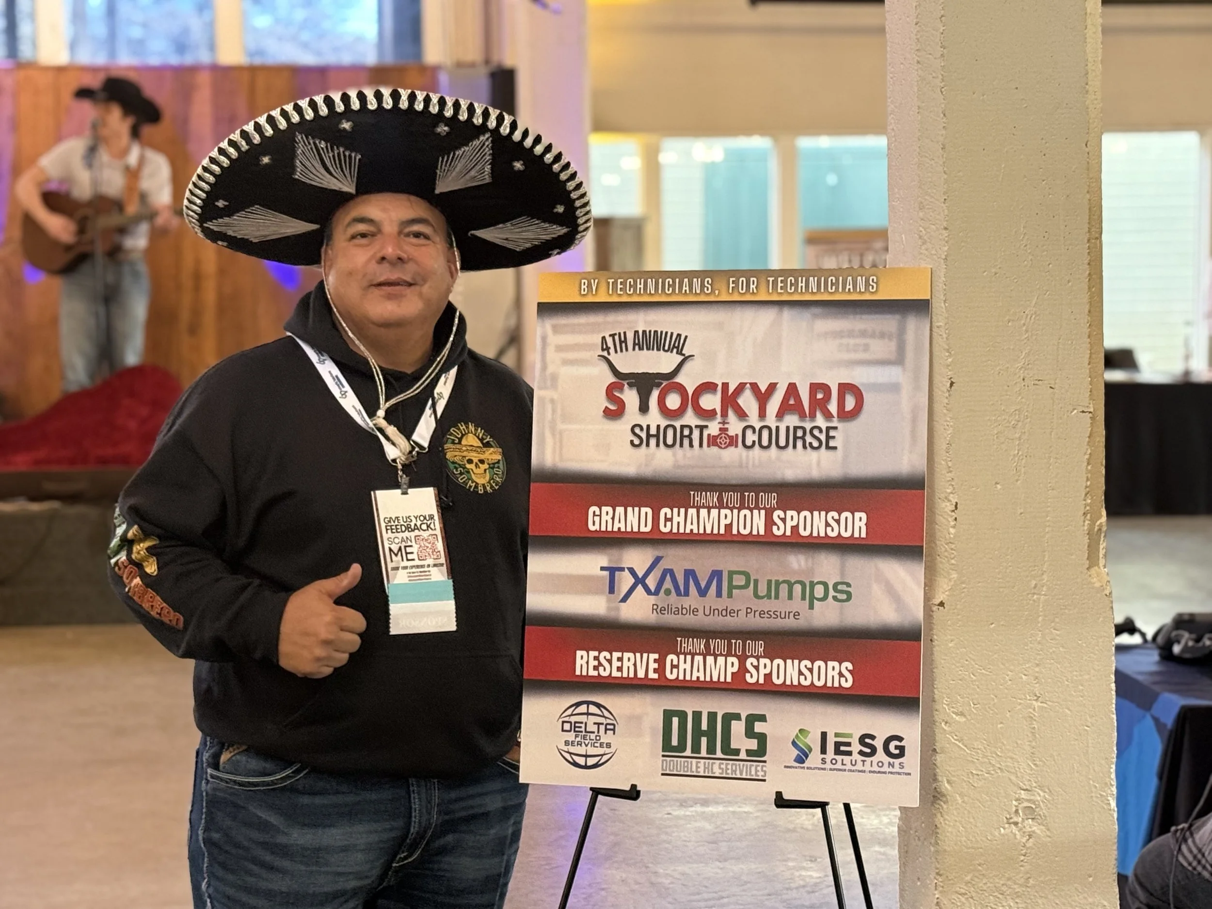Man wearing a large sombrero hat with a black hoodie, standing next to a sponsorship sign at an event. In the background, there's a person playing guitar on stage.