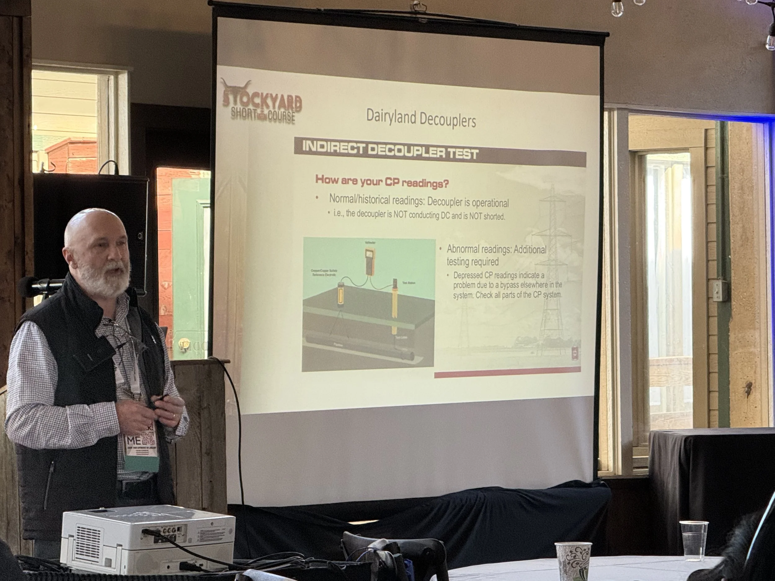 A man with a beard and a bald head giving a presentation at a conference. He is standing in front of a projected slide titled 'Dairyland Decouplers,' which discusses indirect decoupler testing. The slide contains text, a diagram of electrical testing