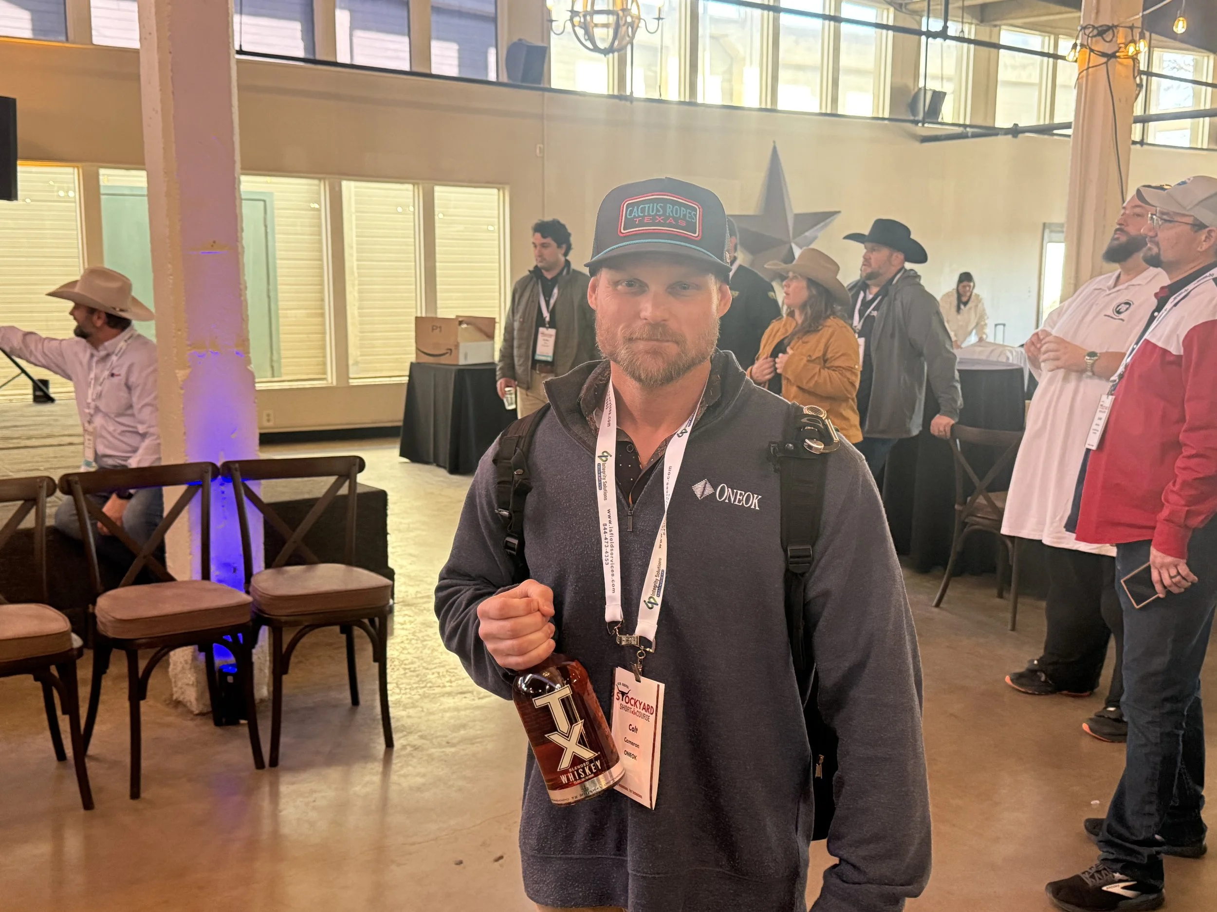 A man with a beard wearing a black cap, a gray jacket, and a backpack is holding a bottle of whiskey and looking at the camera. He is at a conference or event with other attendees in the background.