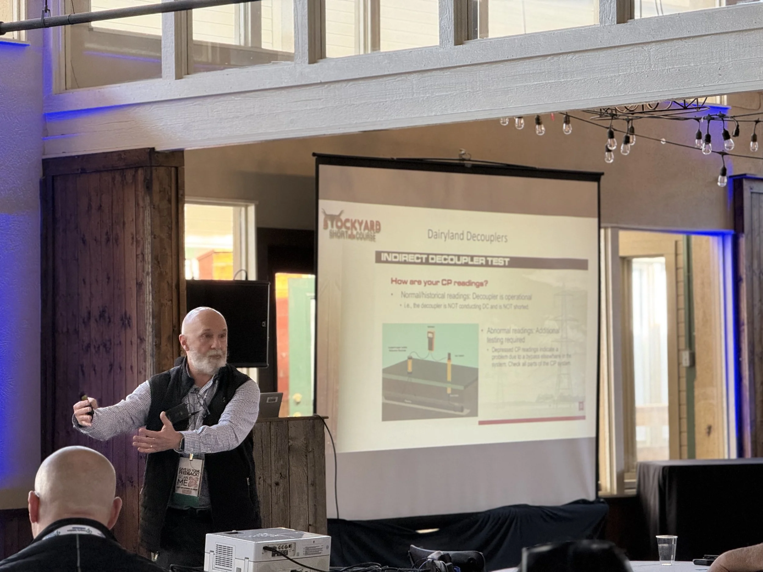 A man presenting at a conference, standing next to a projection screen displaying a slide about Dairyland Decouplers, with an audience seated in front.