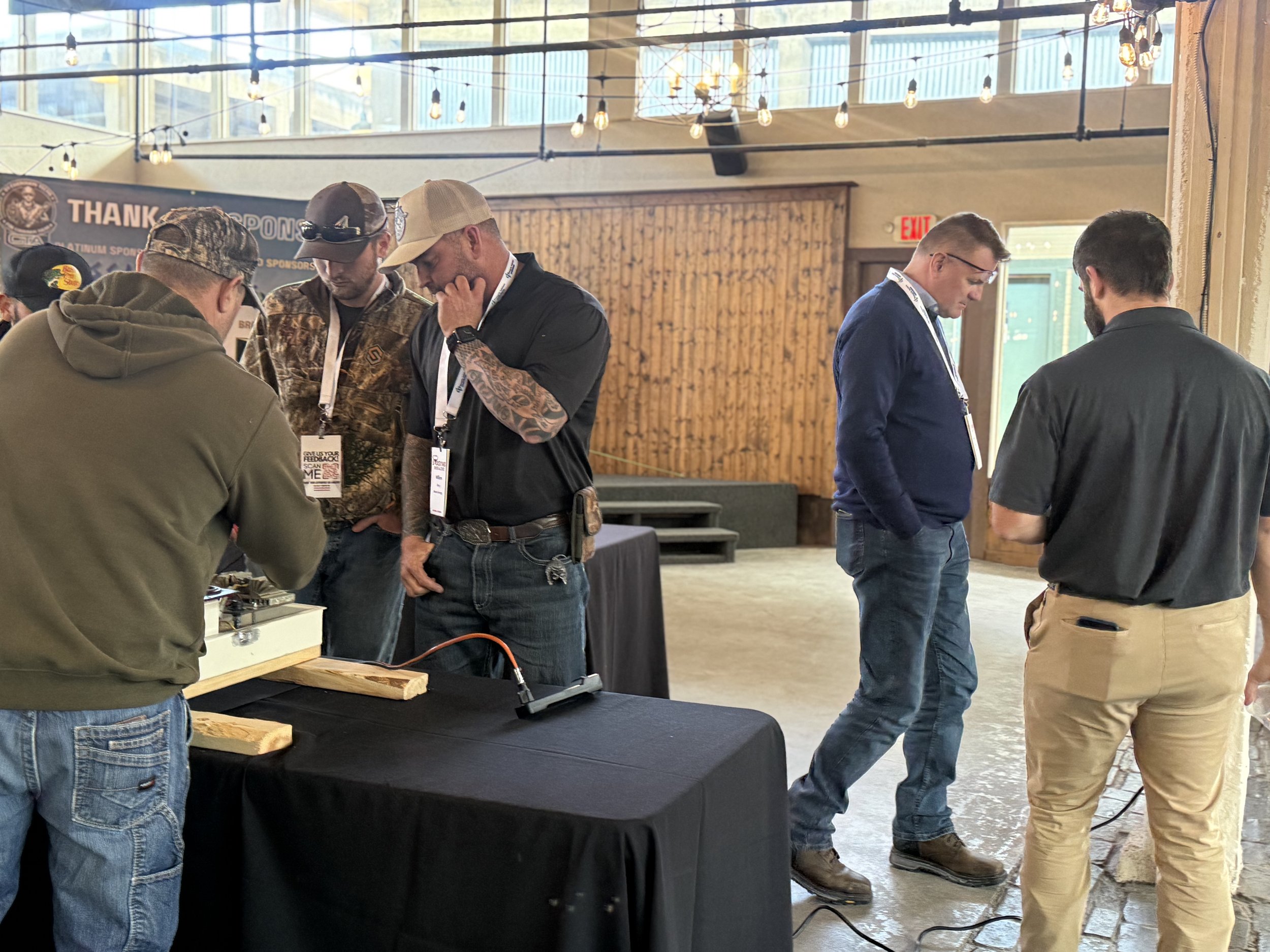Group of men standing around a table at an indoor event, some looking at objects on the table, with warm lighting and wooden decor in the background.