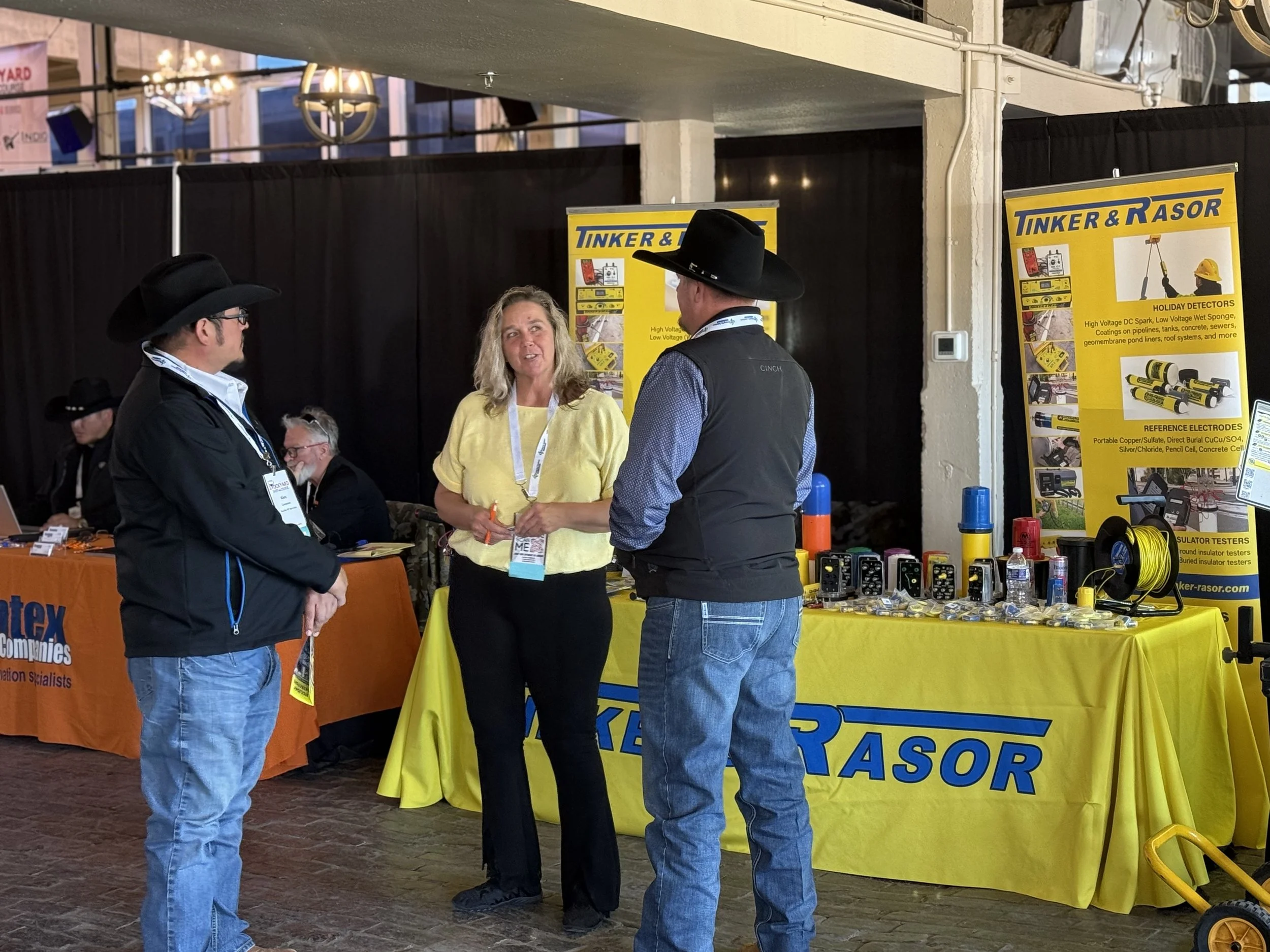 Three people in cowboy hats and casual clothing are standing and talking at an indoor trade show booth with yellow and black banners labeled "Tinker & RASOR," displaying testing equipment on a yellow tablecloth.