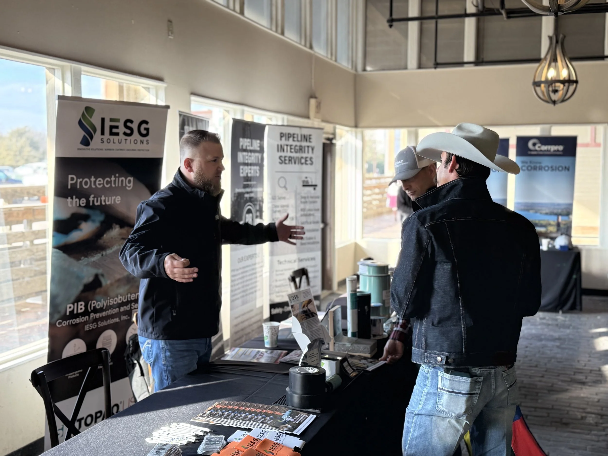 A man with a beard and black jacket is talking to two men in cowboy hats and denim jackets at a trade show booth. The booth has banners for IESG Solutions and related services, with brochures and items on the table.