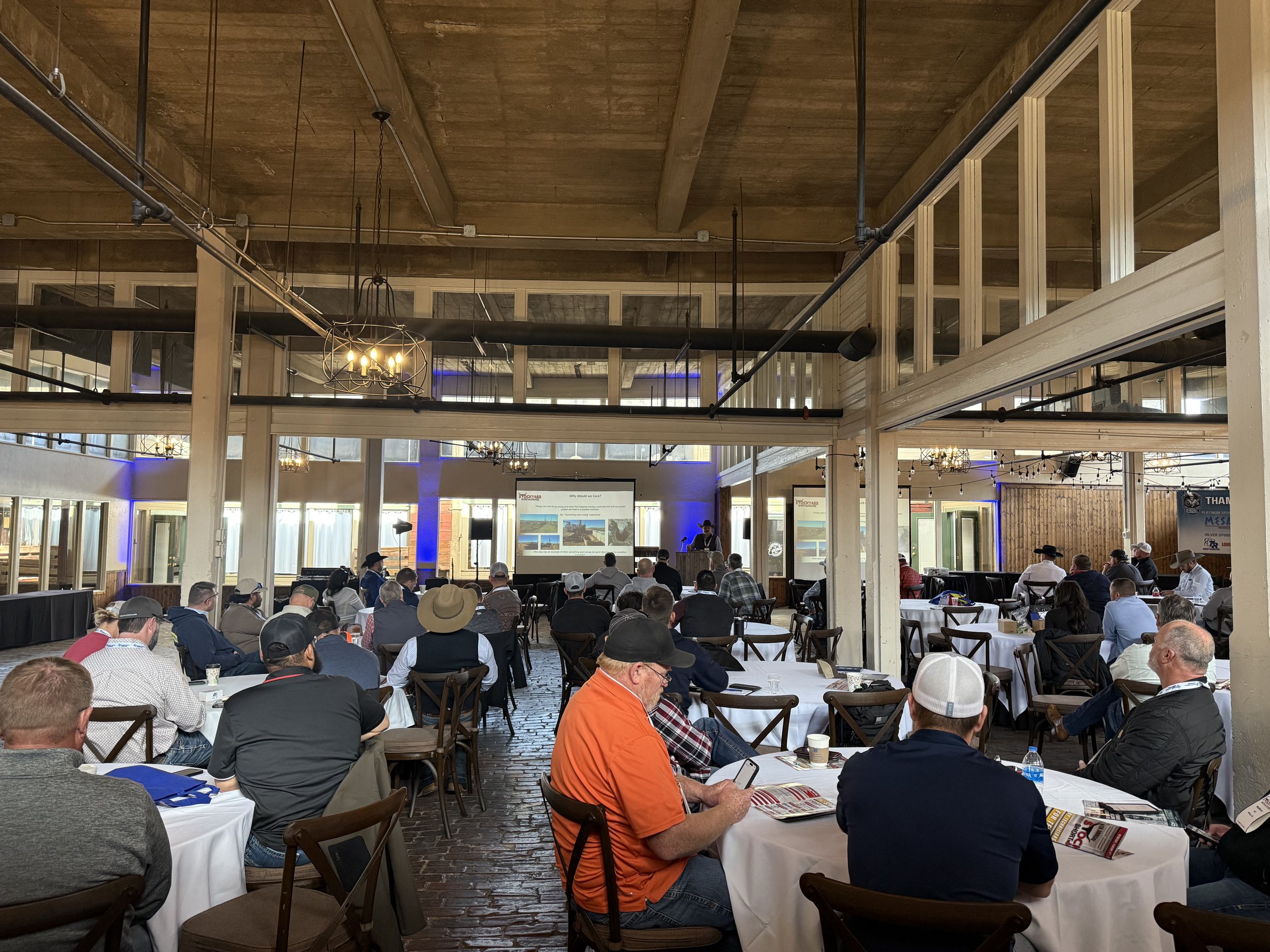 A conference room with people seated at round tables listening to a speaker presentation, with large windows, wooden beams, and decorative chandeliers.