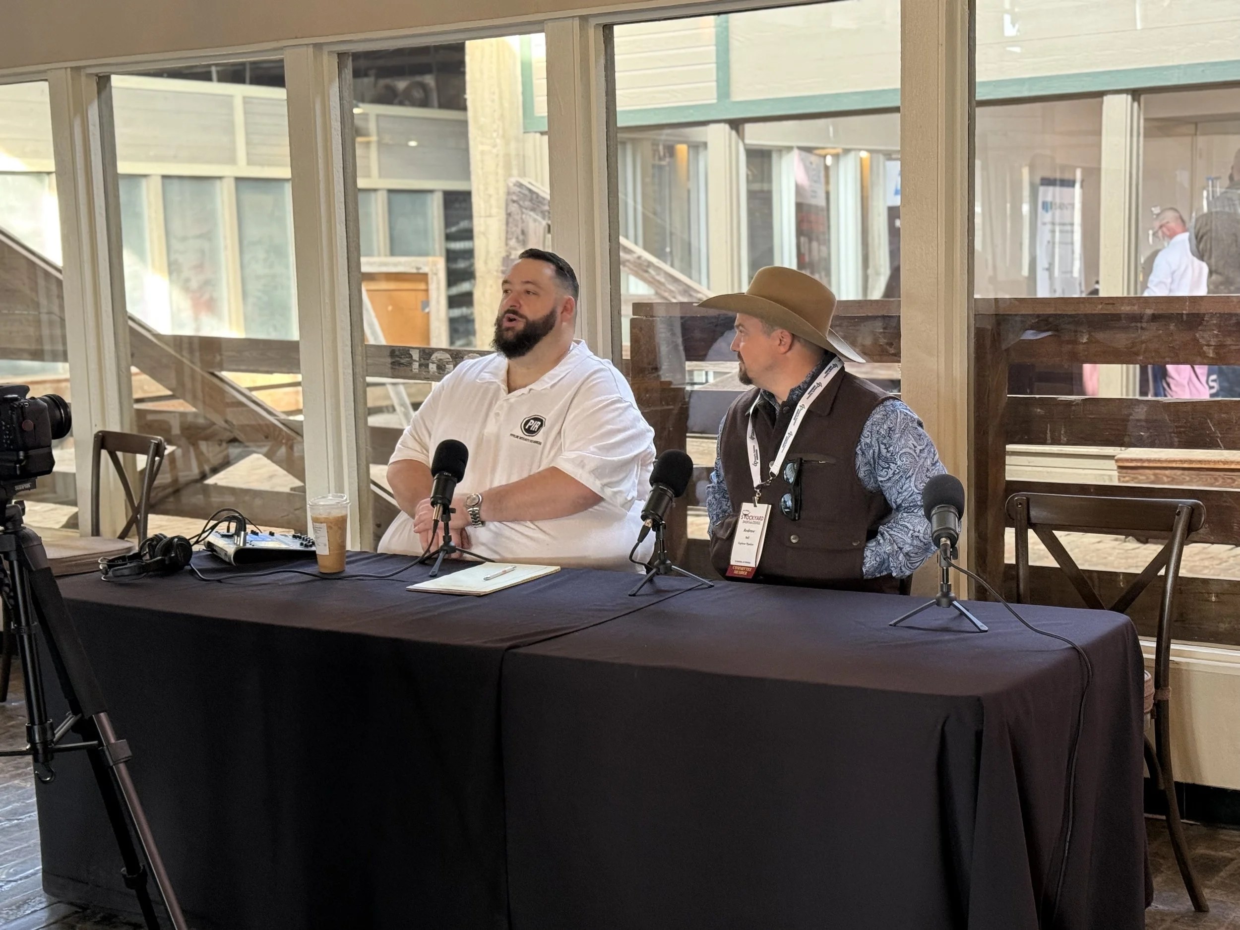 Two men seated at a black table with microphones during a panel discussion or interview at a conference, with a windowed background.