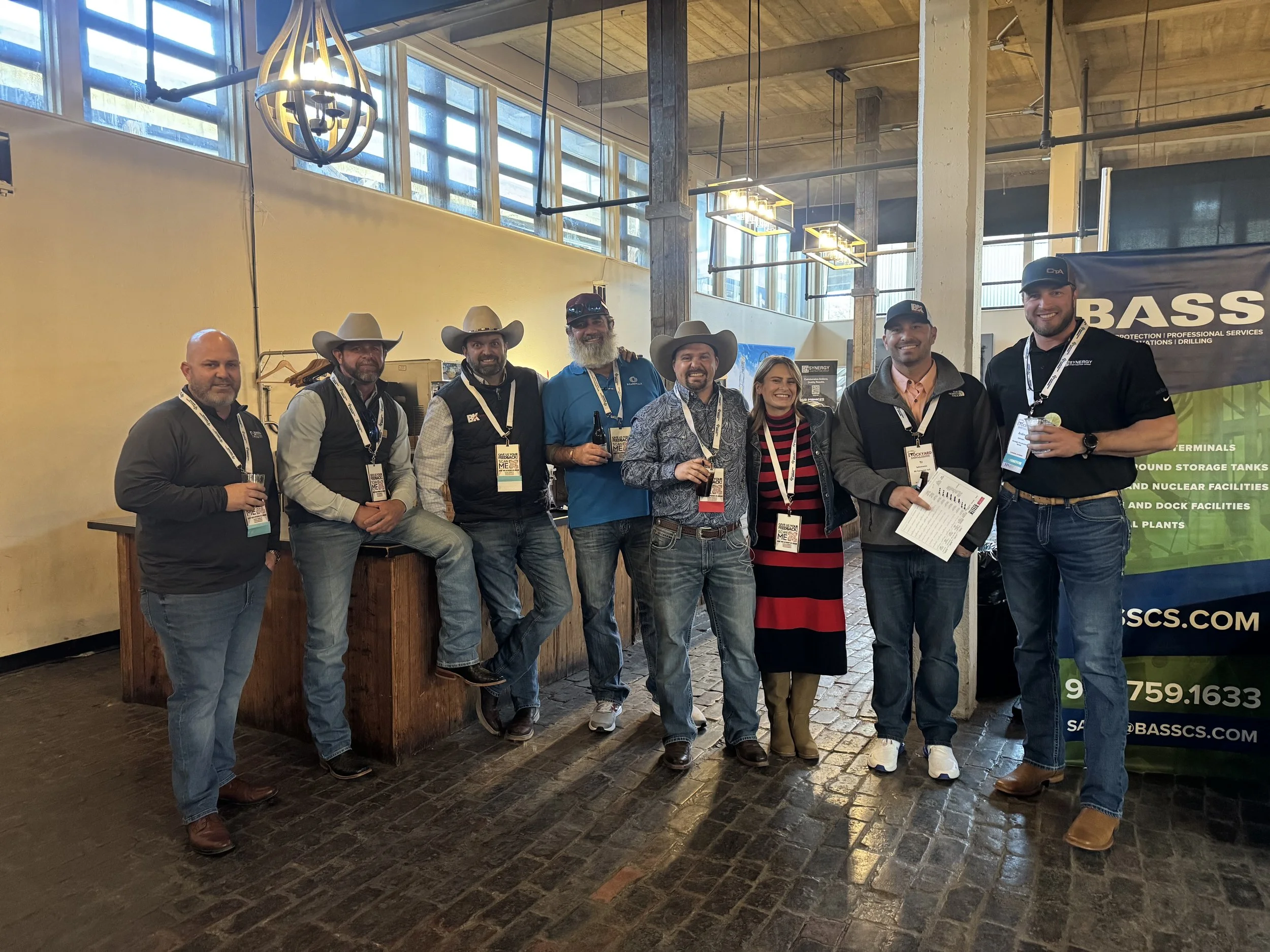 Group of eight people, seven men and one woman, standing together at an indoor event, some wearing cowboy hats, holding drinks, with a banner for BASCS in the background.